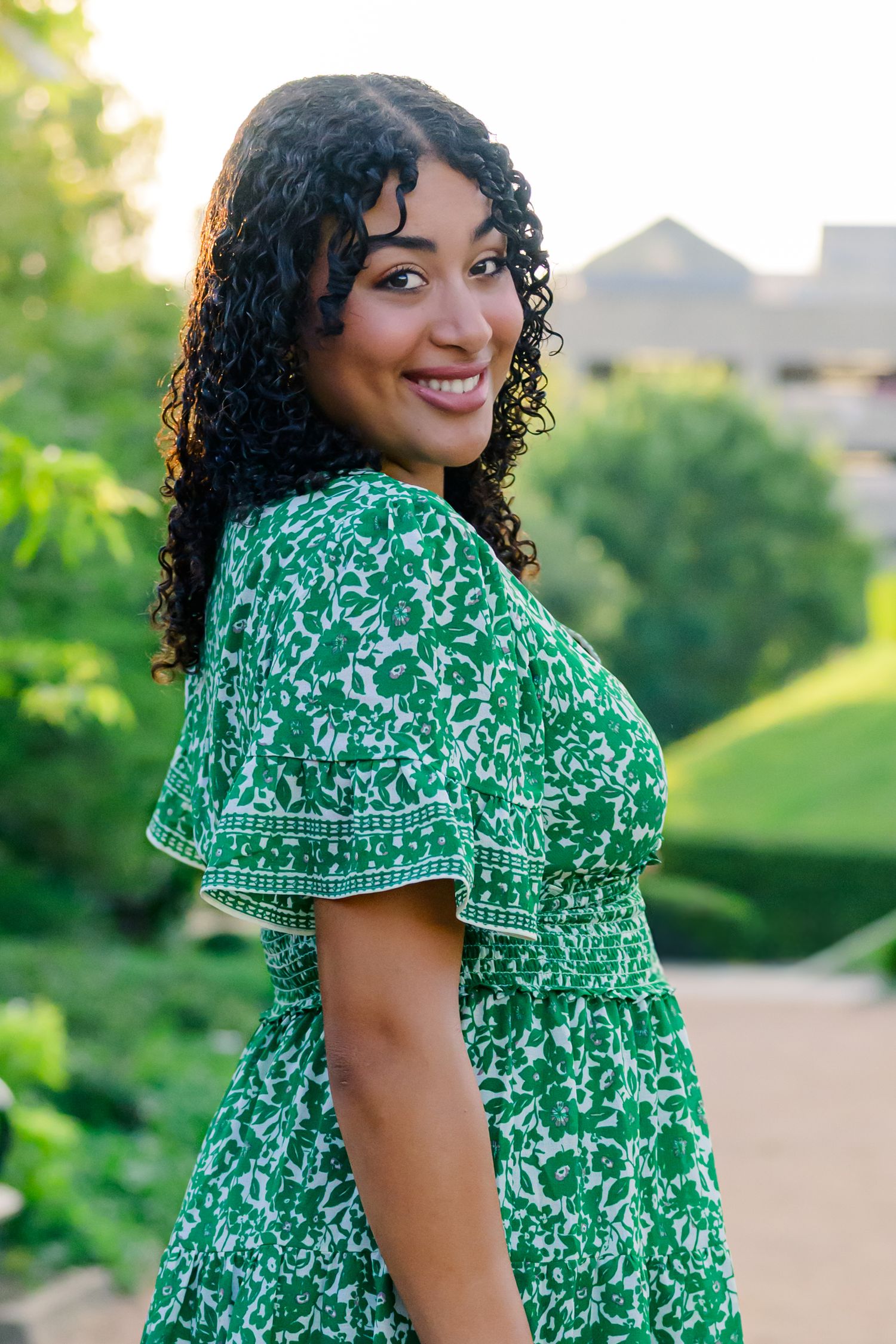 A person in a green floral dress sits on a wooden bench holding a black hat while smiling outdoors.