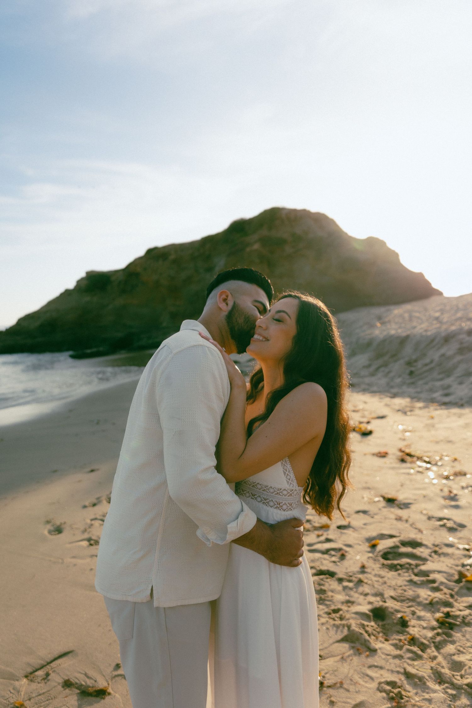A romantic couple in white attire embraces on a sandy beach with cliffs and ocean in the background at sunset.