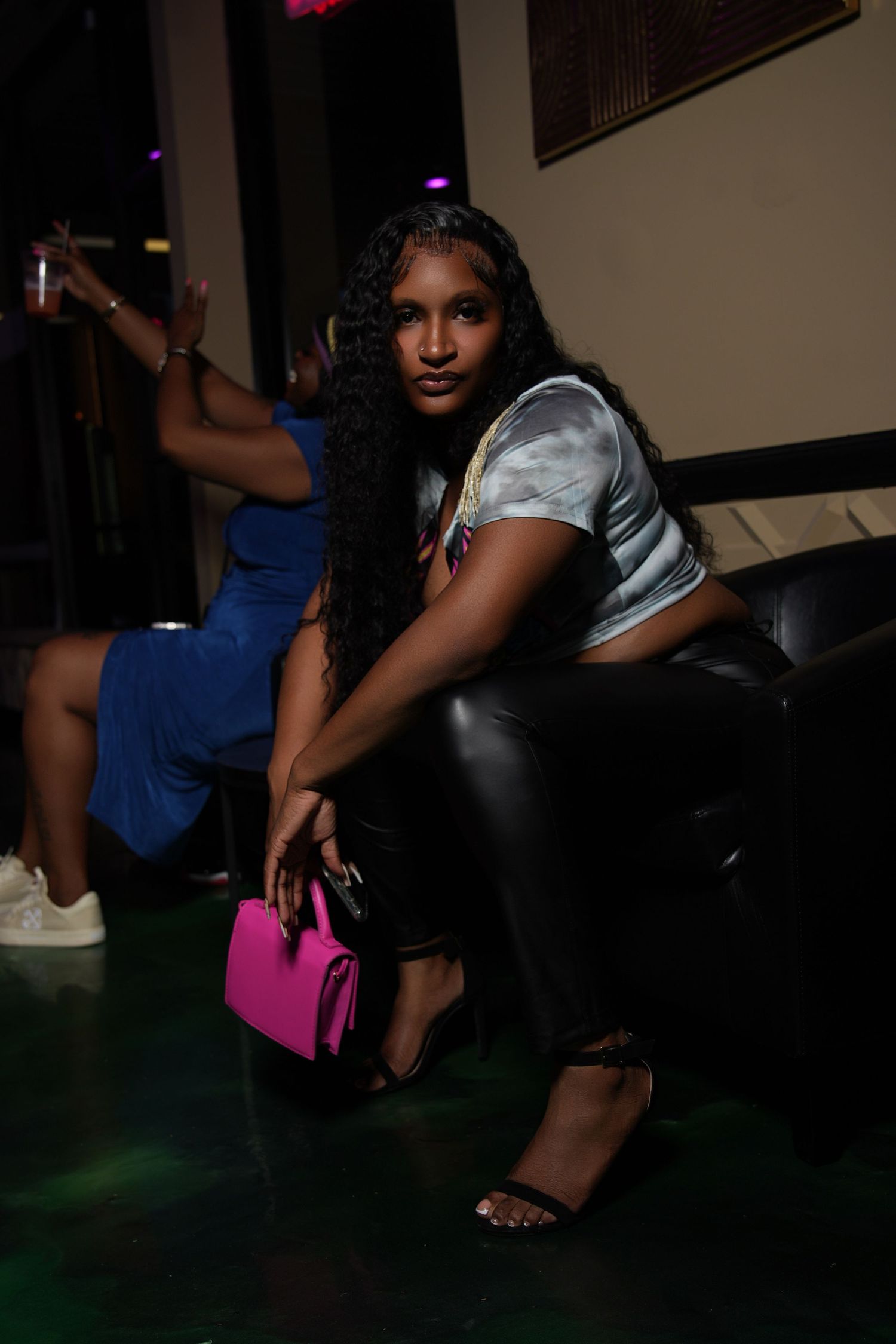 A person in a white top and dark pants poses confidently in a dimly lit space with a bright pink handbag.