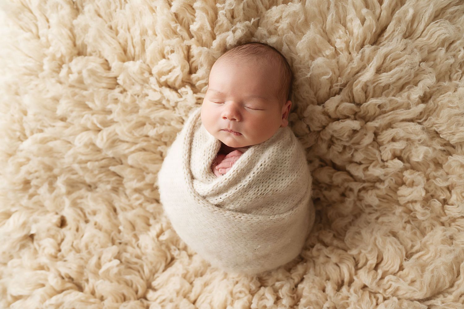Newborn baby swaddled in white wrap sleeping peacefully on fluffy cream rug.
