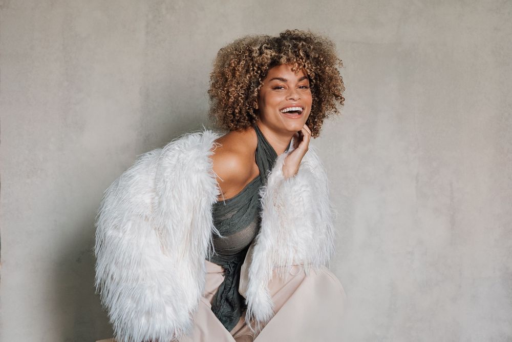 Person in white furry jacket laughing joyfully against a neutral background.
