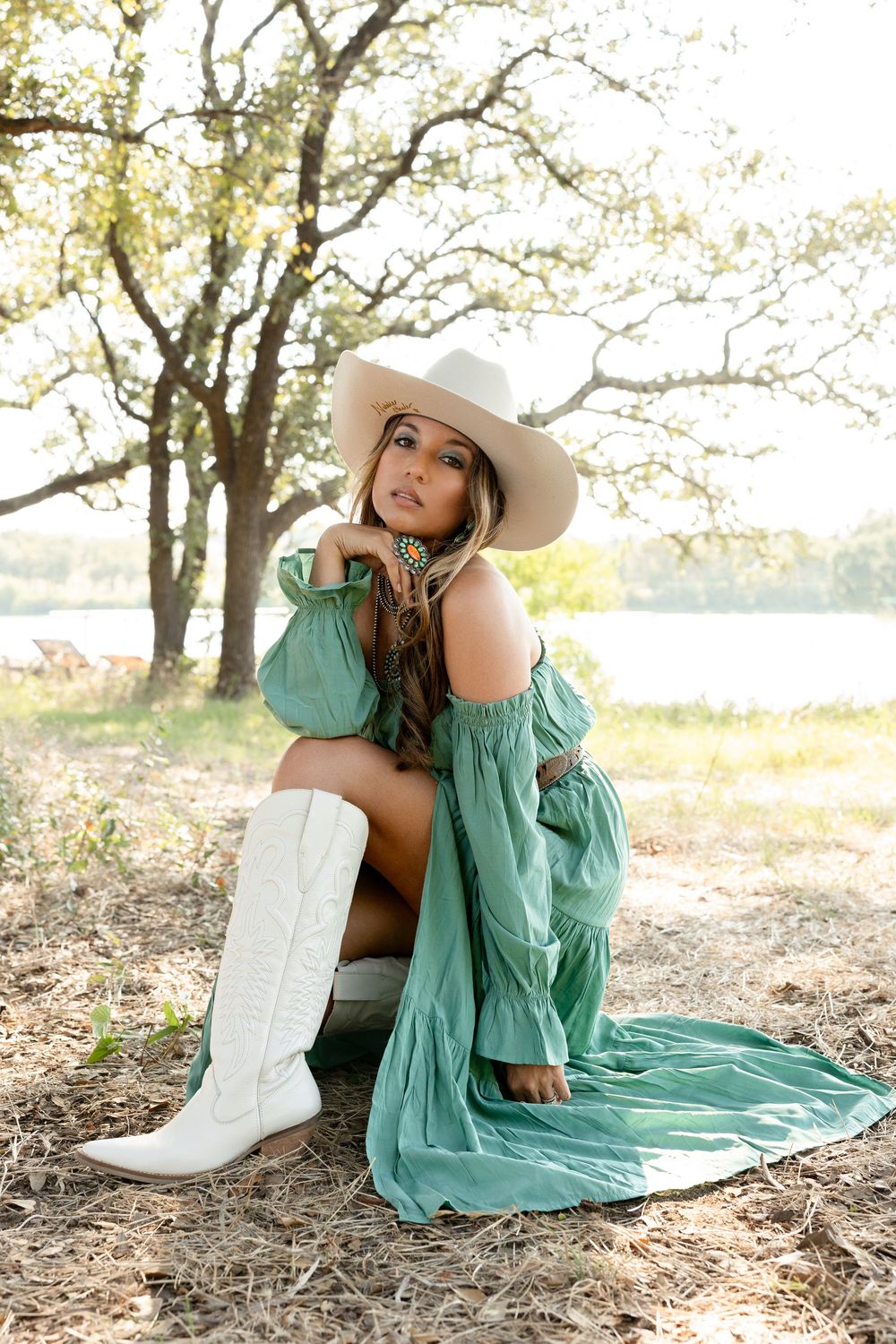 Woman in mint green dress and white cowboy boots poses gracefully on ground with sunlit trees in background.
