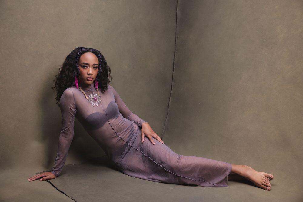 Fashion model in lilac dress poses gracefully against textured brown backdrop for editorial photoshoot.
