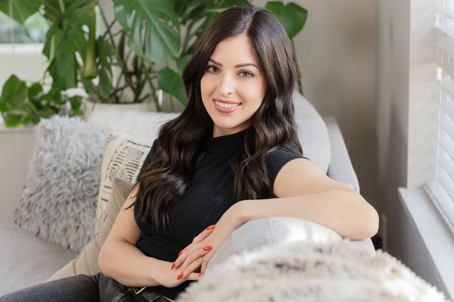 Portrait of brunette sitting on couch near houseplants in bright interior setting.