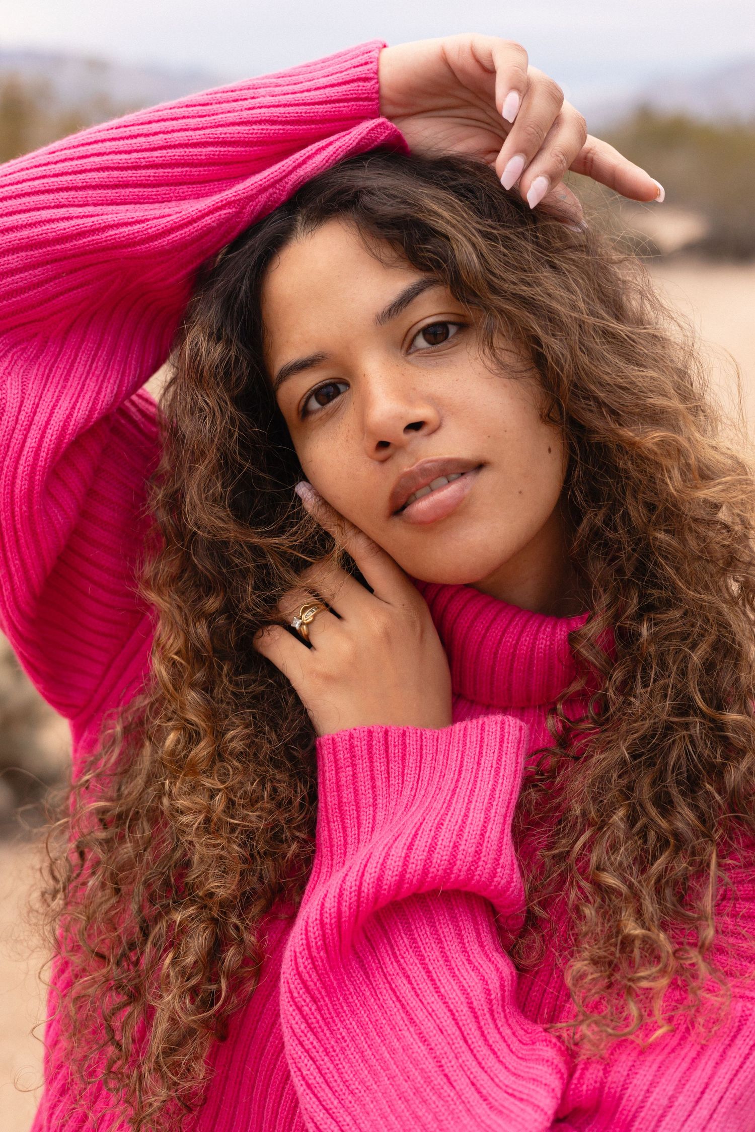 Model wearing bright pink ribbed sweater with long curly hair posing outdoors in natural light.