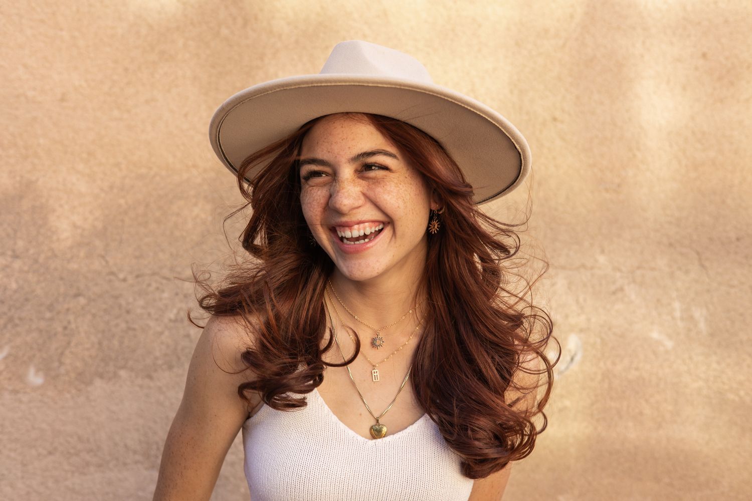 Happy woman in white top and beige hat laughing against textured wall in warm sunlight.