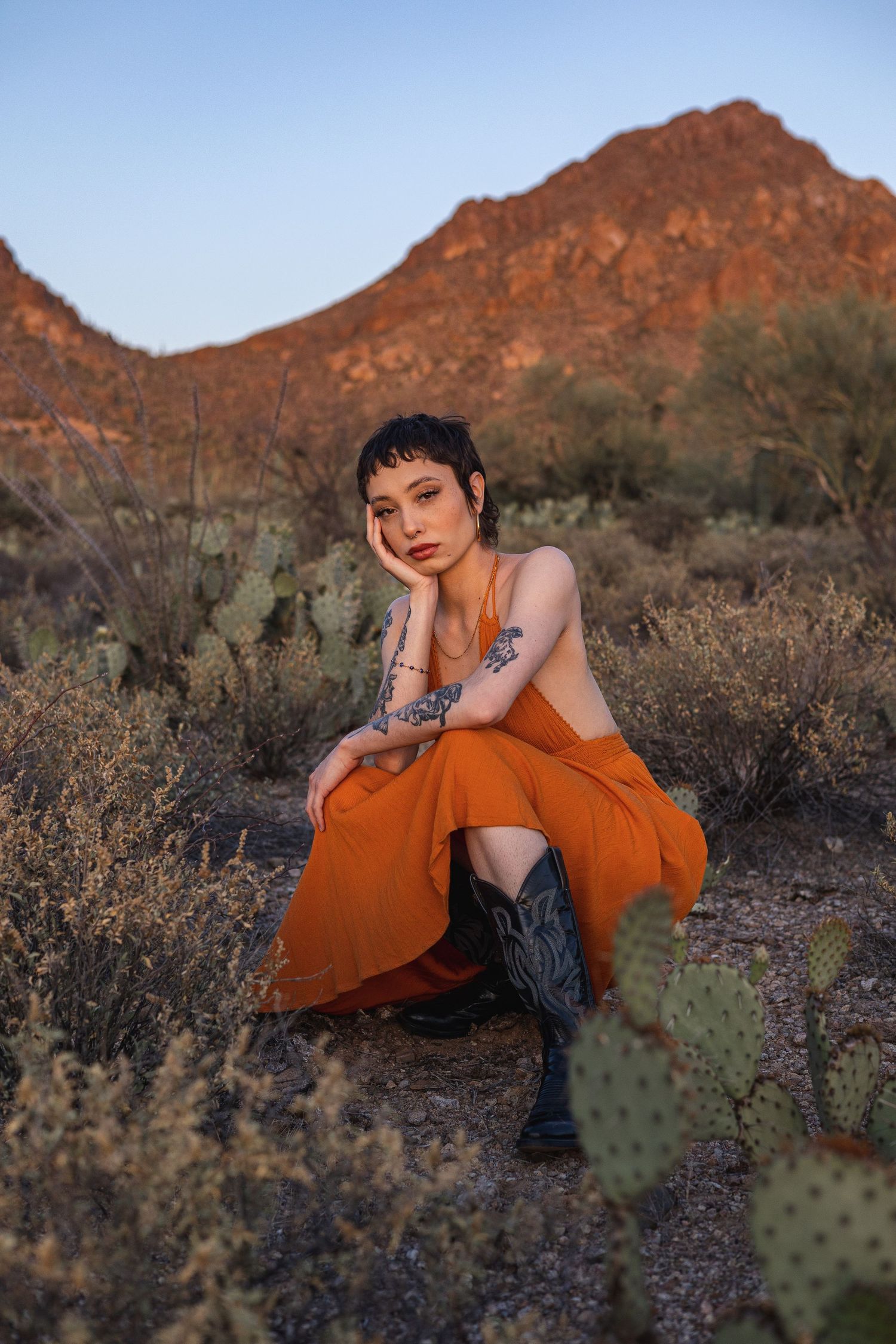 A portrait in golden desert light shows someone in an orange dress and boots sitting among cacti with red mountains behind.