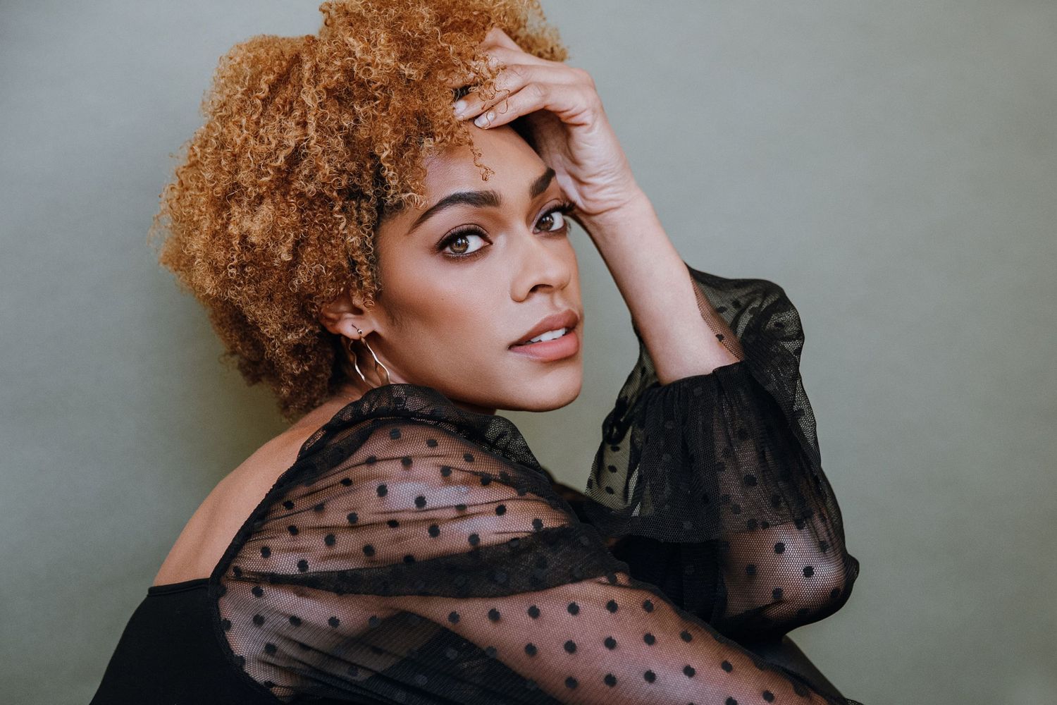 Woman with curly auburn hair wearing a black sheer polka dot off-shoulder top, posing elegantly against a green background.