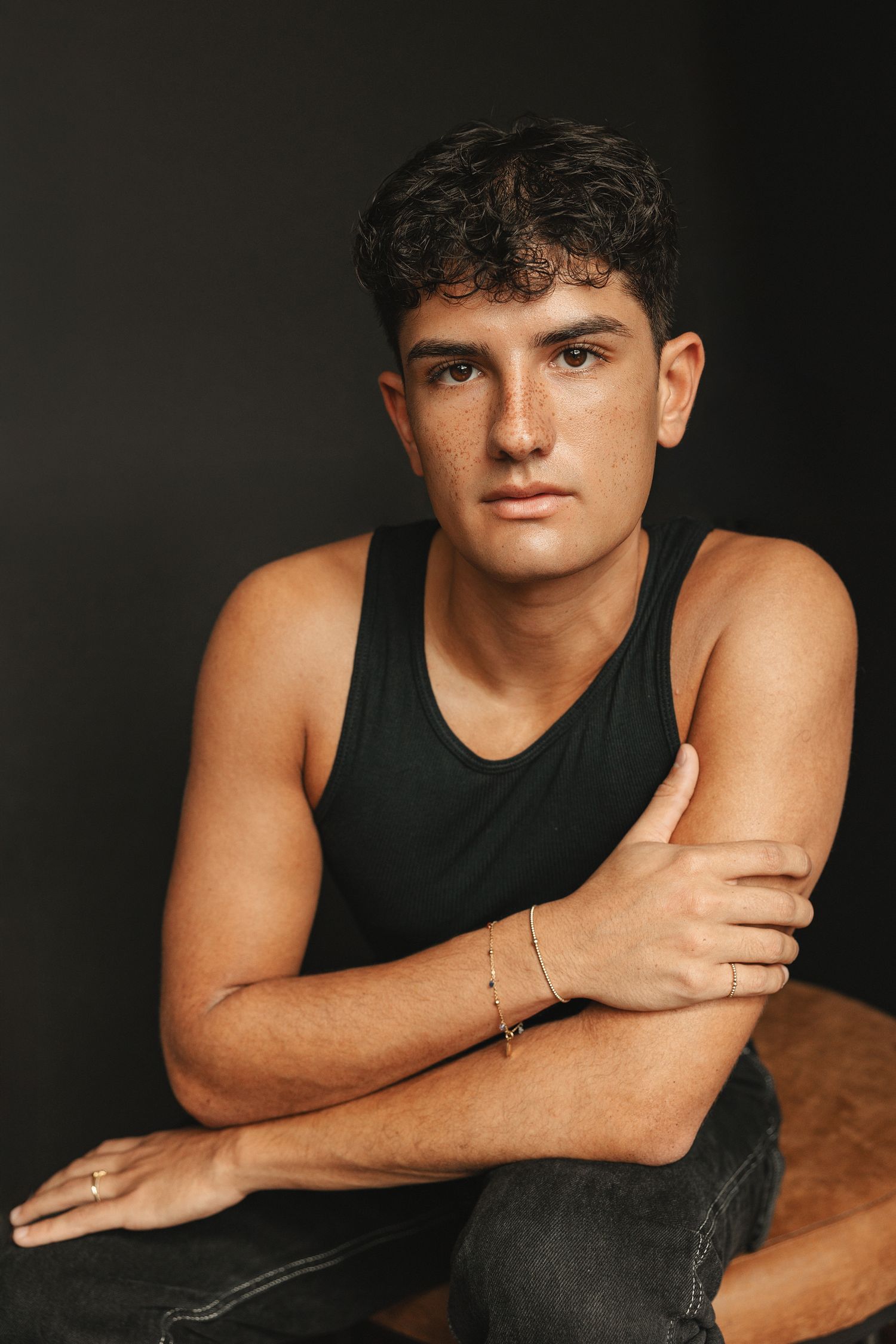 Young man in black tank top with gold bracelet poses confidently against dark background.