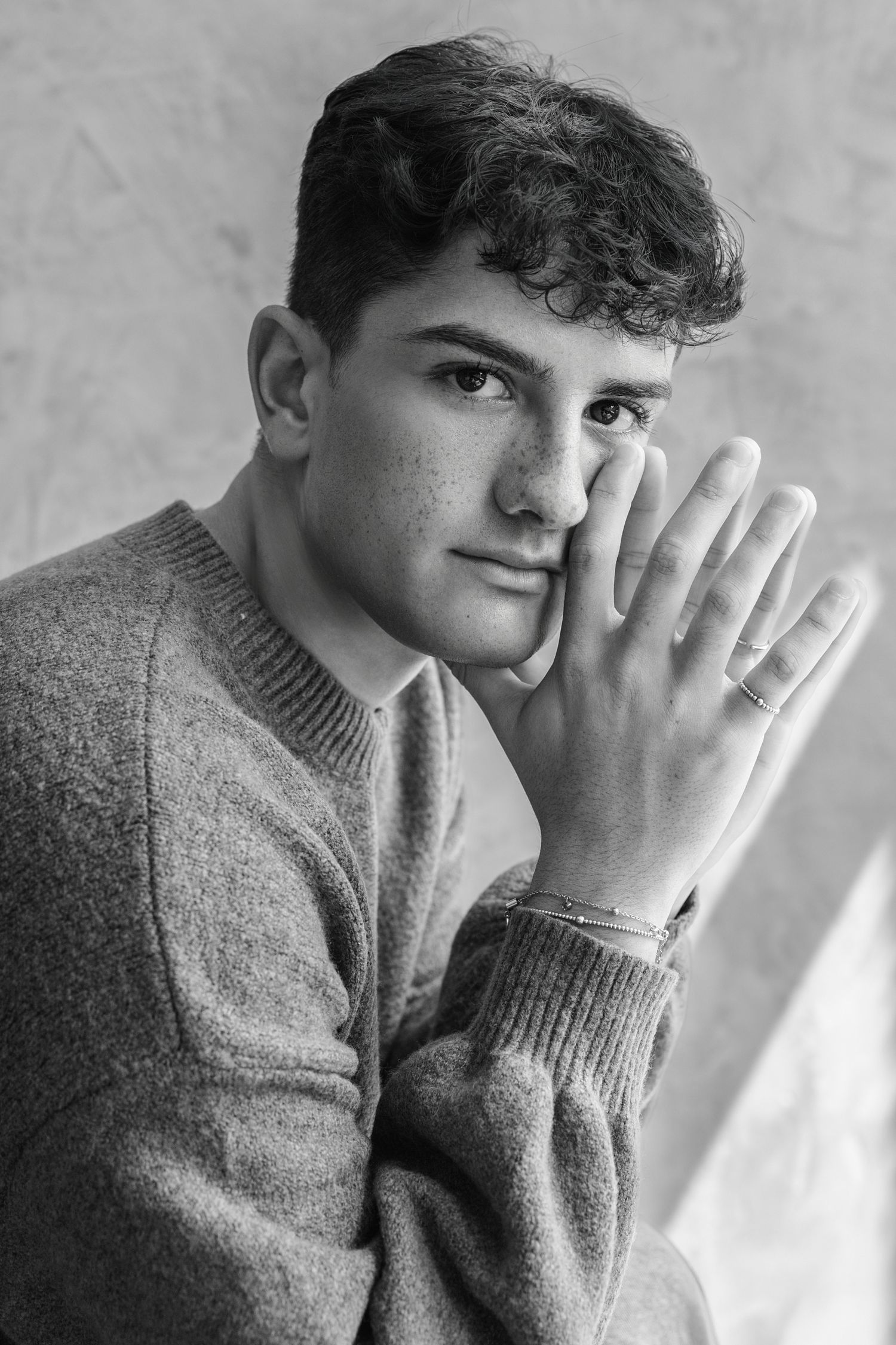 Black and white portrait of a young man in a sweater resting his face on his hand, wearing a bracelet.