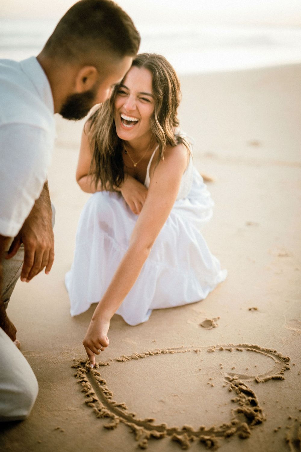 A joyful couple in white clothing draws a heart in the sand on a beach at sunset during their romantic photoshoot.
