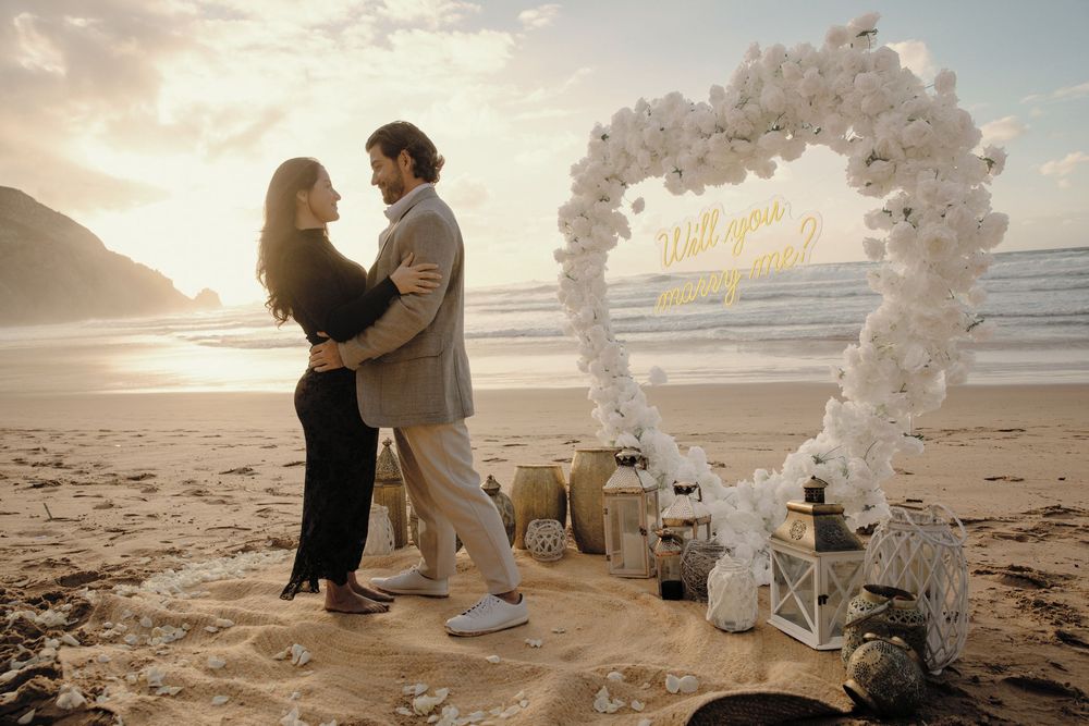 Romantic beach wedding ceremony setup with white floral arches and decor along the sandy shoreline at sunset.