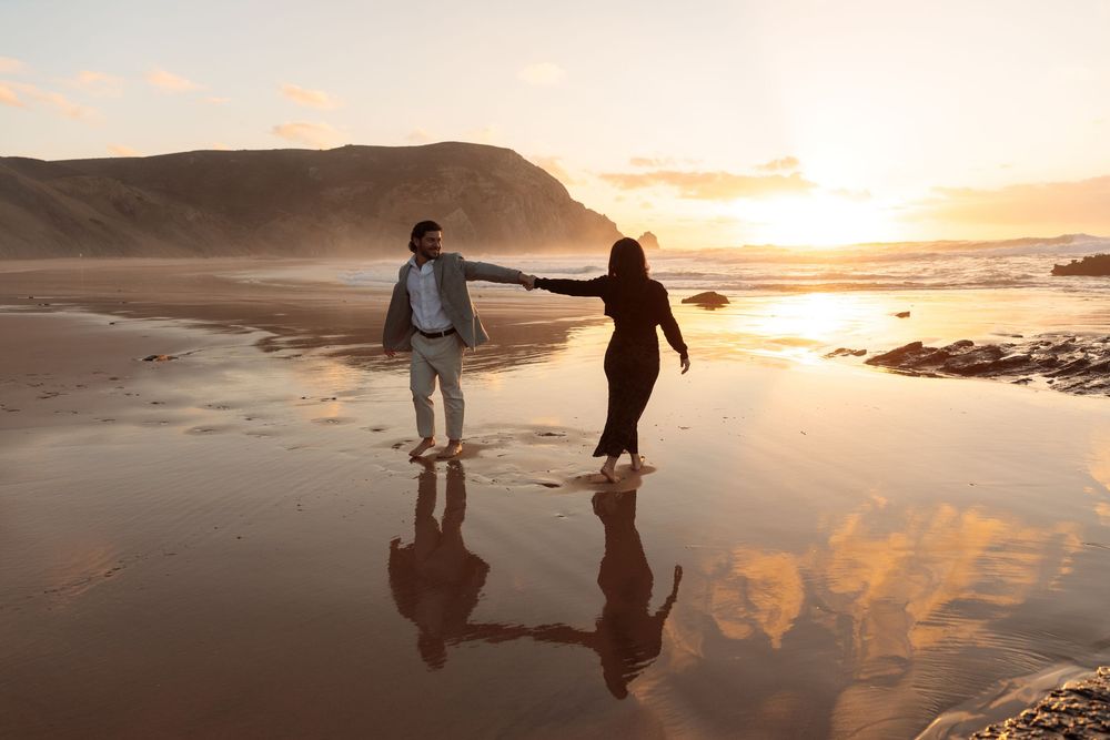 Two silhouettes walk along a reflective beach at sunset with golden light illuminating the sky and water.