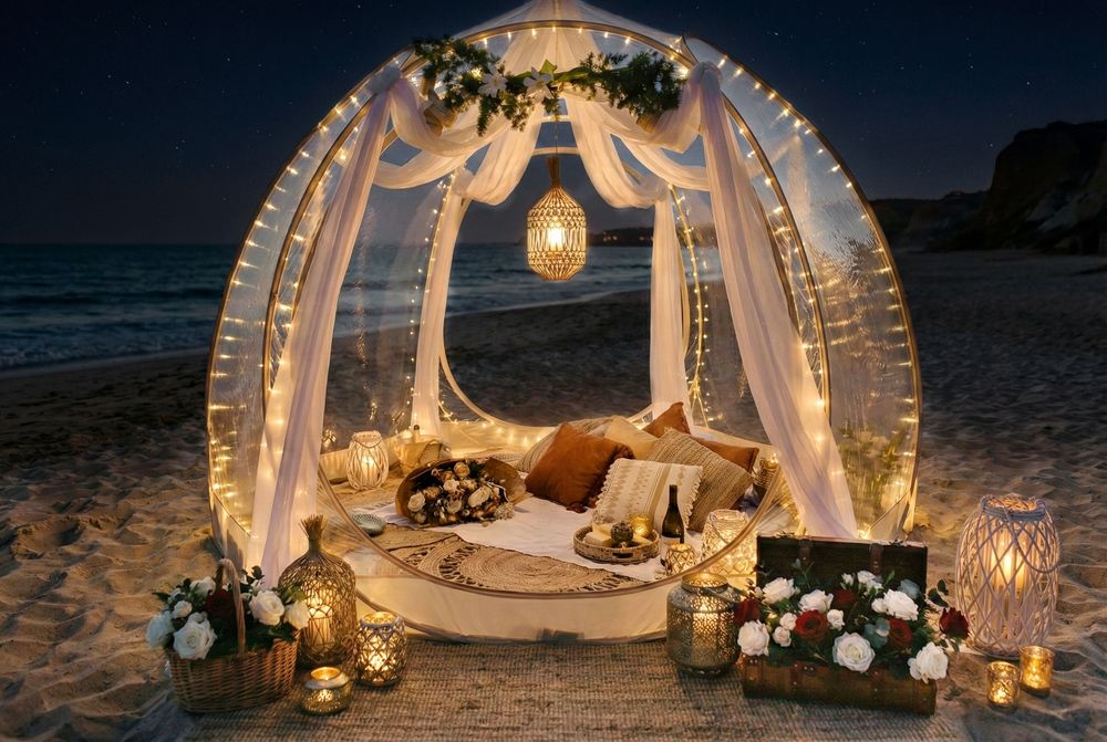 A romantic beach dinner setup with white drapes, lanterns, and candles creating an intimate atmosphere at sunset.