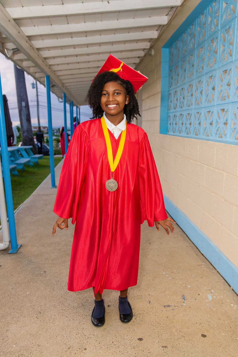 Graduate wearing bright red cap and gown with gold honors cord and medal stands in covered walkway by blue wall.