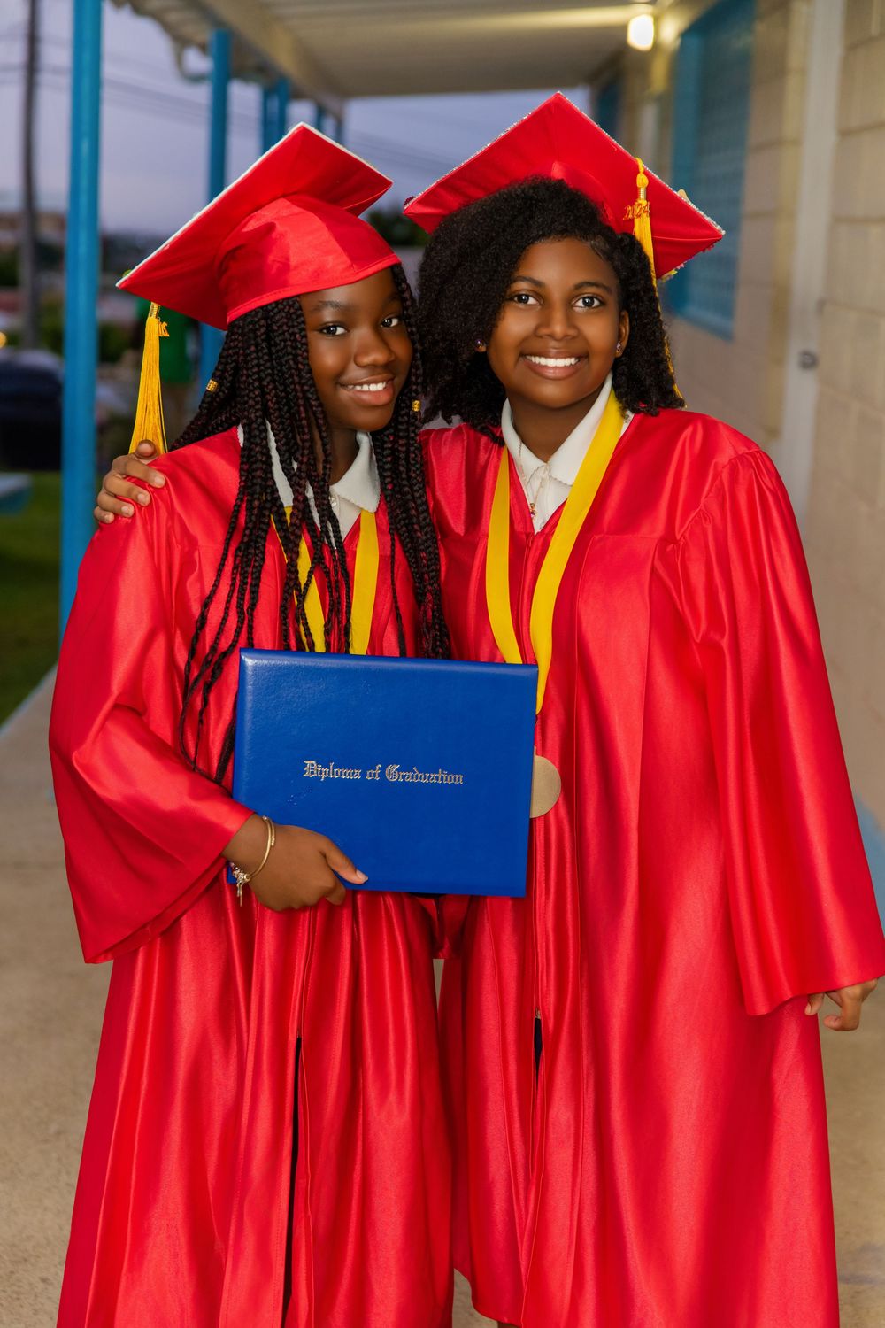 Two graduates in bright red caps and gowns stand together holding a blue diploma cover with gold accents and tassels.