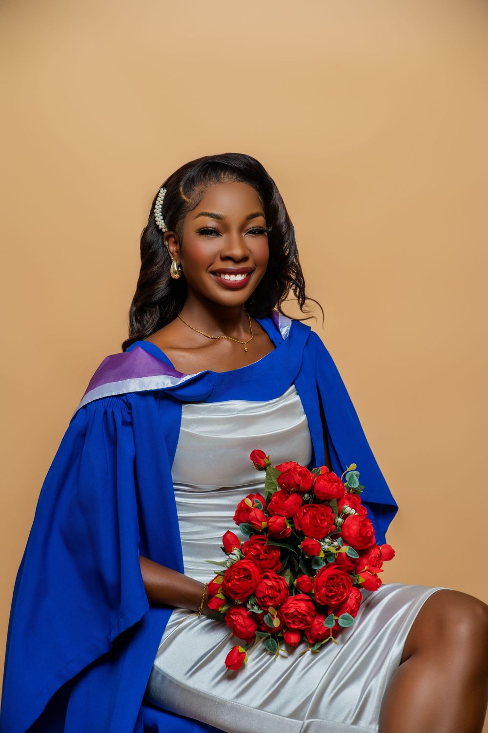 Graduate in blue cap and gown holding vibrant red rose bouquet poses against beige backdrop for graduation photos.