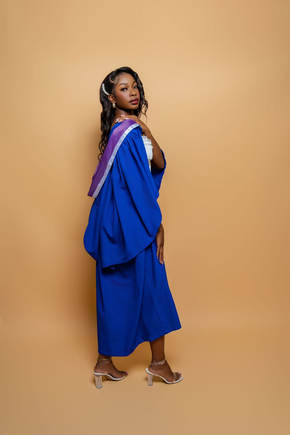 Graduate in royal blue graduation gown and cap poses elegantly against beige background wearing silver heels in Antigua by Mark Blan.