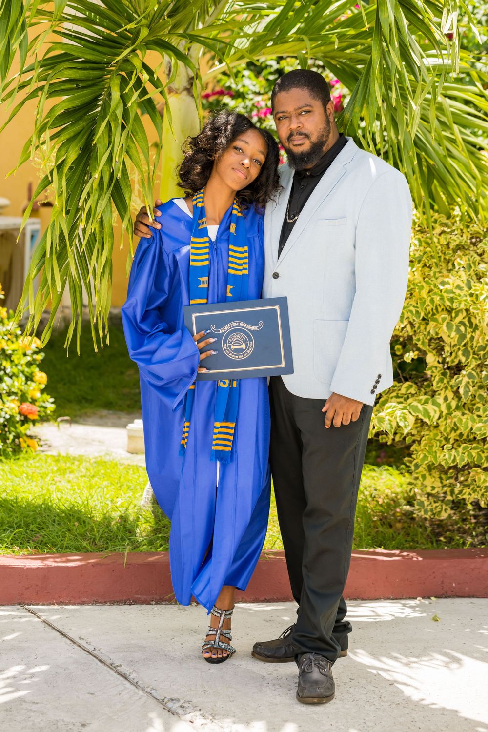 A graduate in royal blue commencement regalia poses with companion in white blazer against tropical foliage background in Antigua.