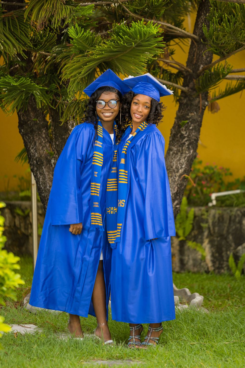 Two graduates in bright royal blue caps, gowns and kente stoles stand together outdoors under palm trees in Antigua by Mark Blan.