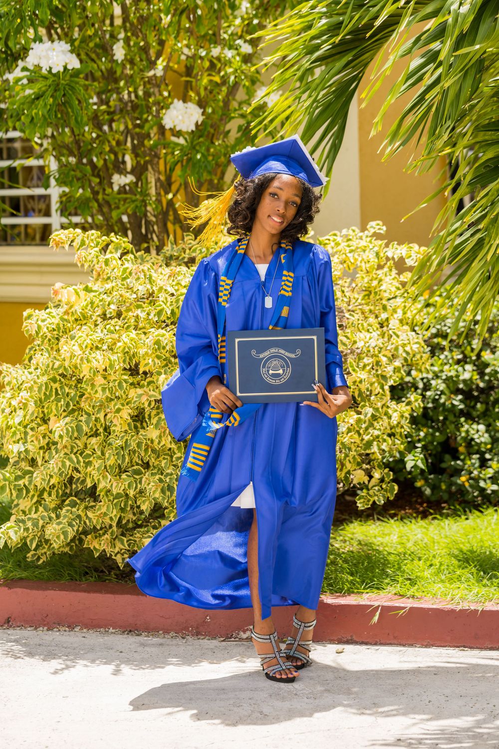 Graduate in bright blue cap and gown holding diploma stands outdoors near tropical plants on sunny day in Antigua by Mark Blan.