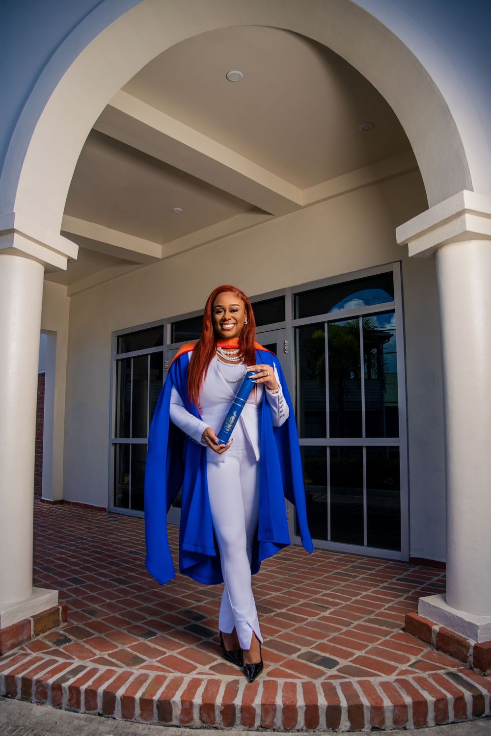 Graduate in blue academic regalia poses with diploma holder on brick entryway steps of building with white columns.