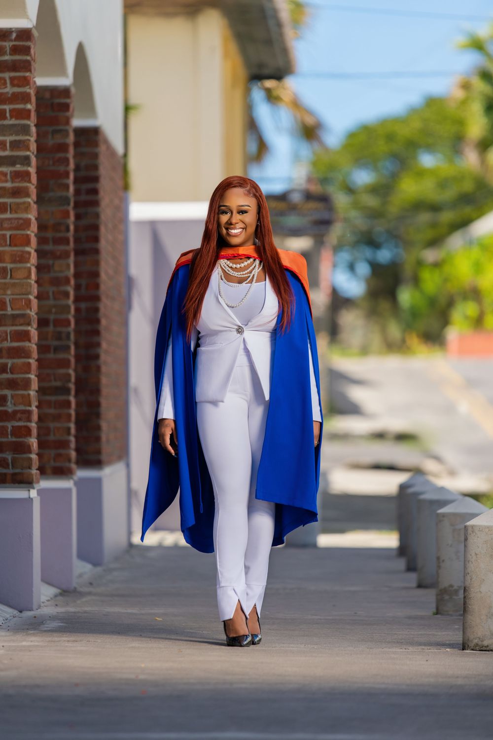A person wearing a chic white pantsuit and bright blue cape coat walks confidently down a brick-lined sidewalk.