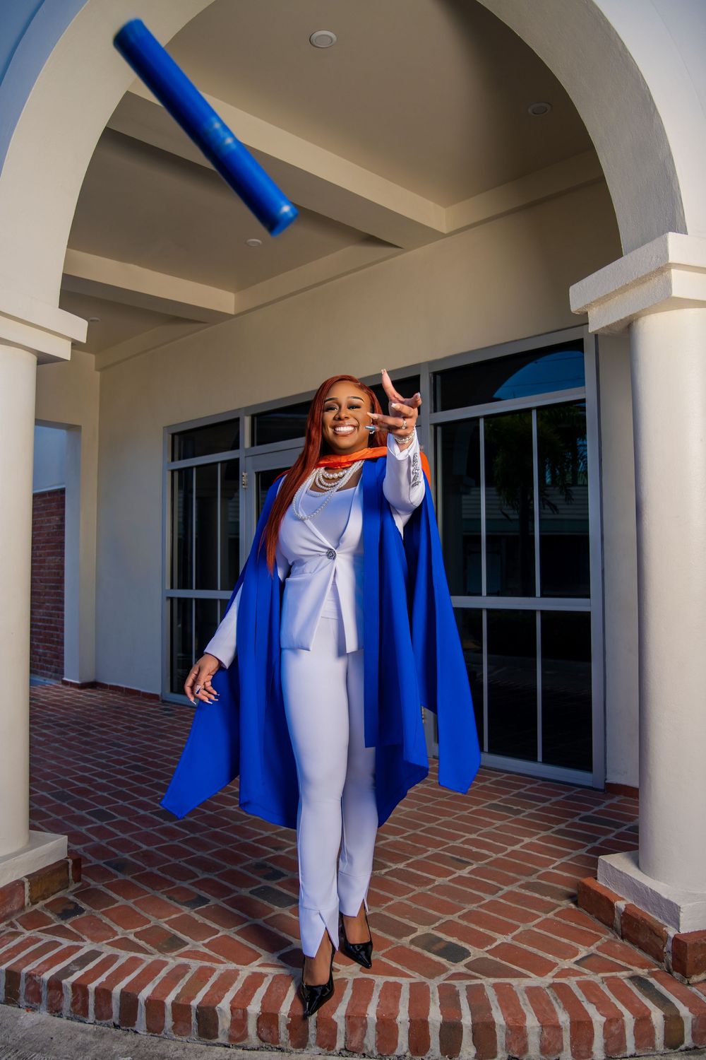 Graduate standing on brick steps in blue graduation gown and white outfit celebrates by tossing a blue diploma holder in the air.