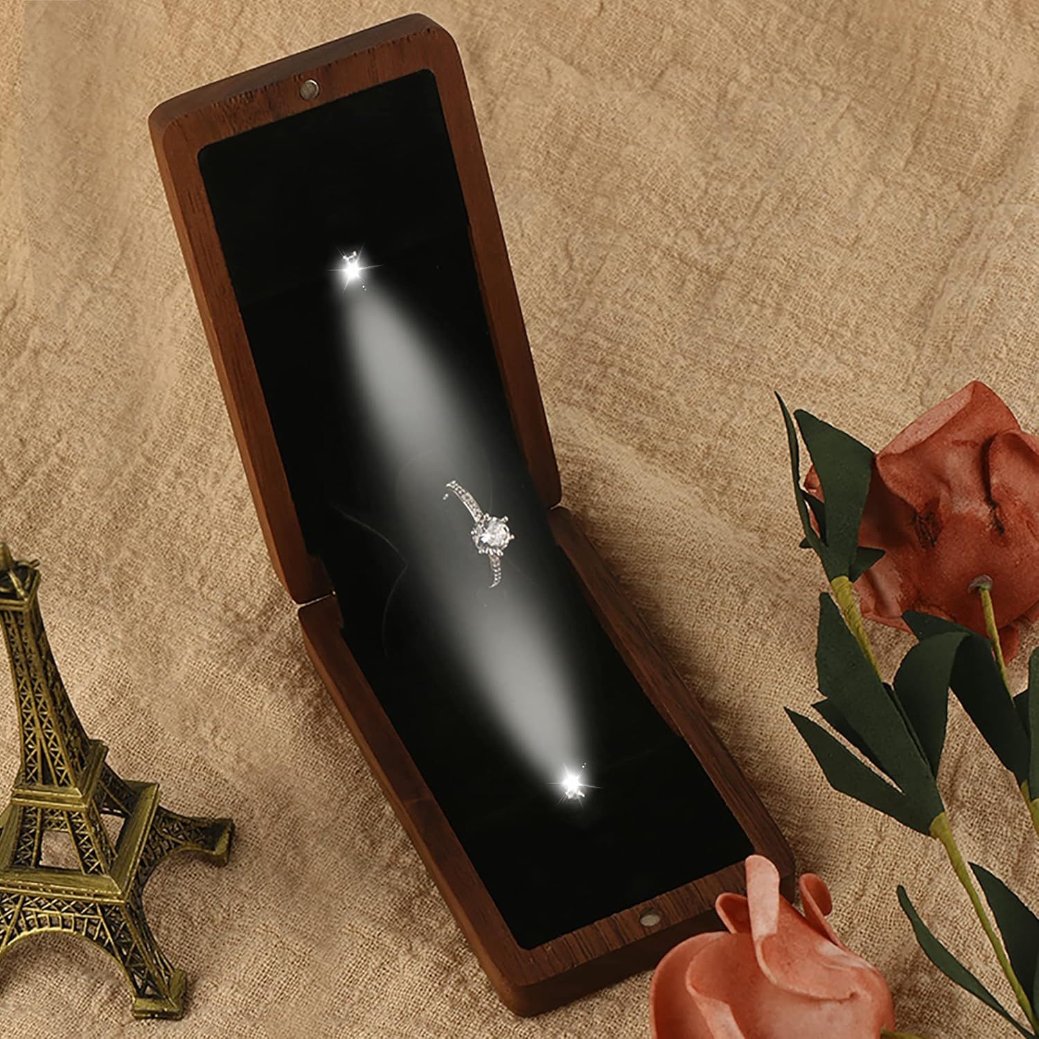 A ring box with a spotlight shining on a diamond ring, displayed next to a mini Eiffel Tower and coral roses.