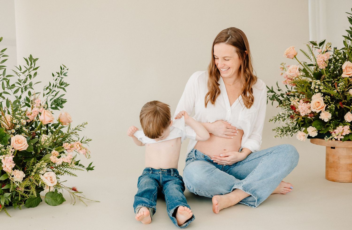 A parent and toddler sit on a white floor next to pink floral arrangements in terracotta pots.