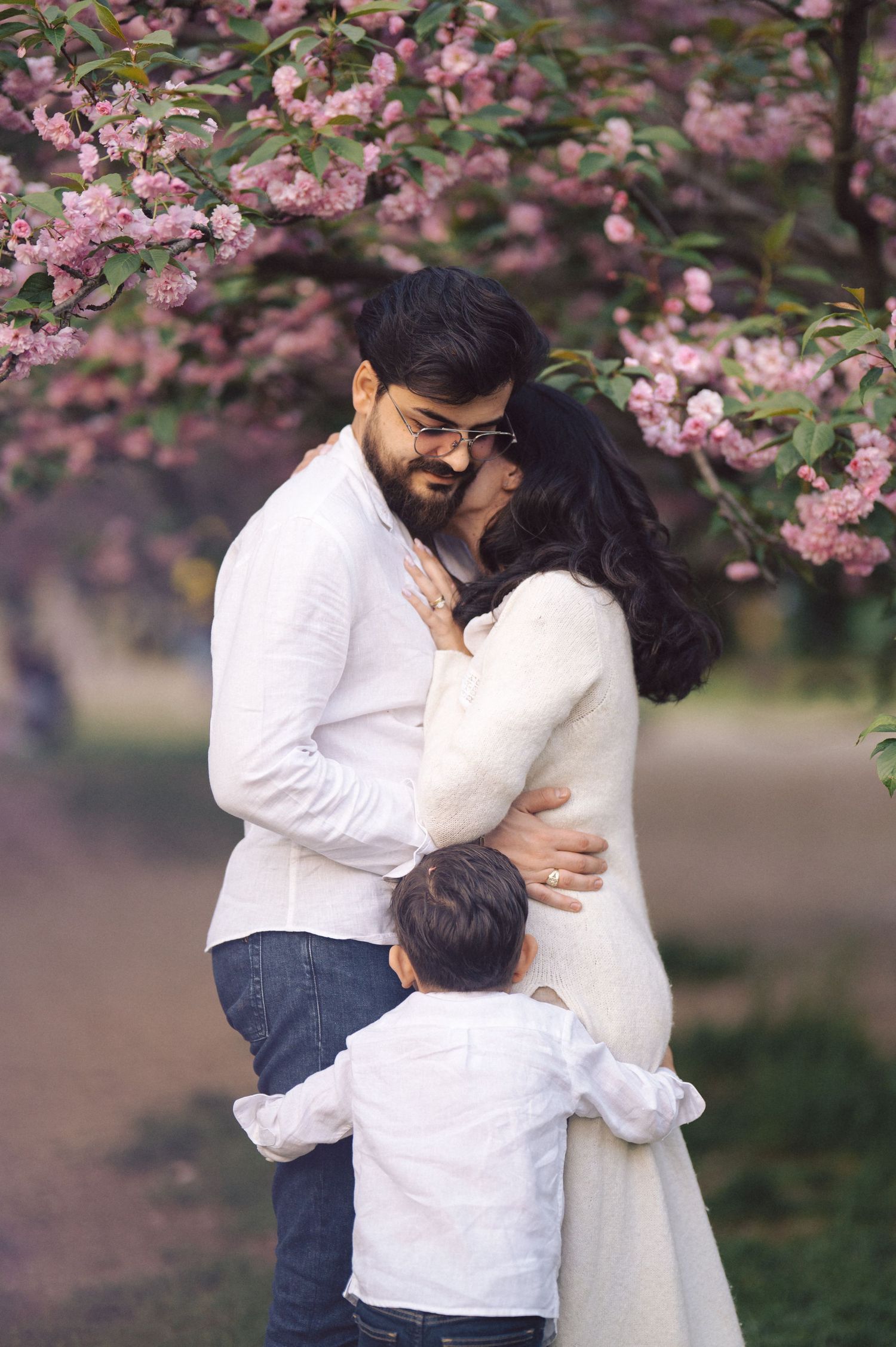 A couple embraces lovingly under blooming pink cherry blossoms while a small child looks on.