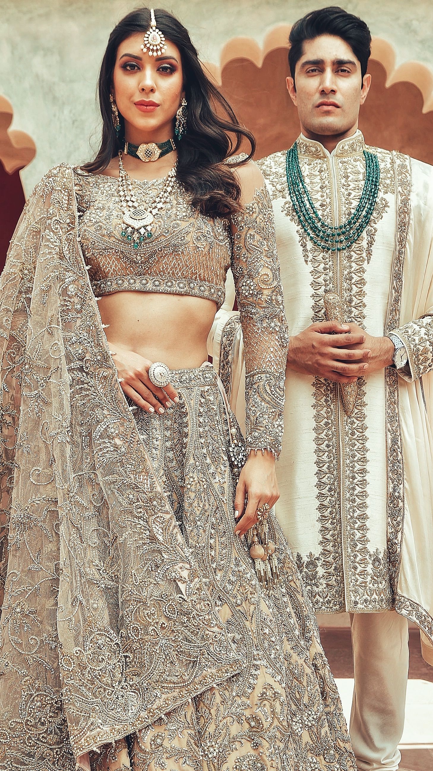 Couple in elegant Indian bridal wear, woman in embroidered lehenga with jewelry, man in cream sherwani with emerald necklace.