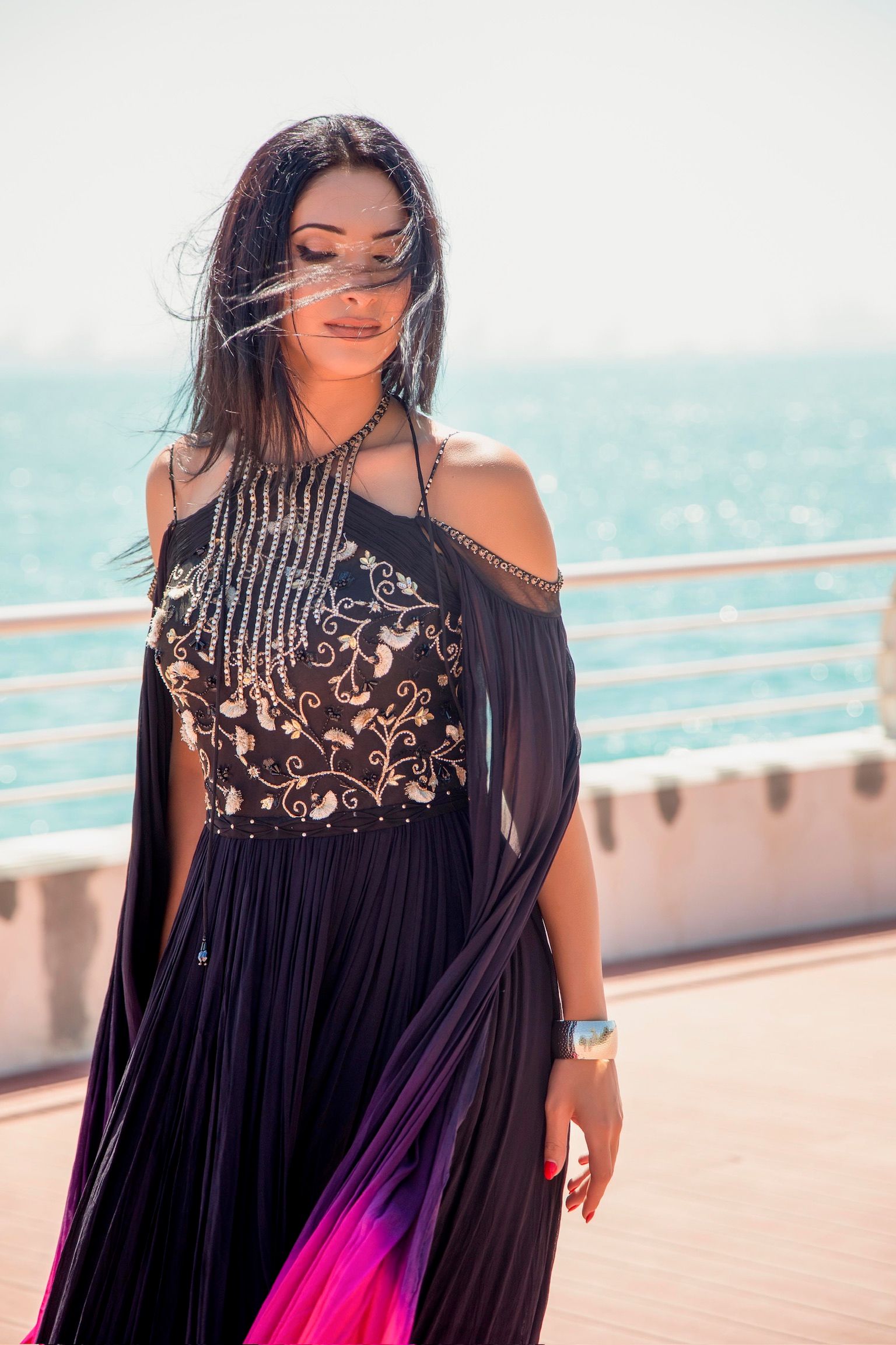 Woman in elegant black embroidered halter gown with pink ombre hem poses on seaside promenade on sunny day.