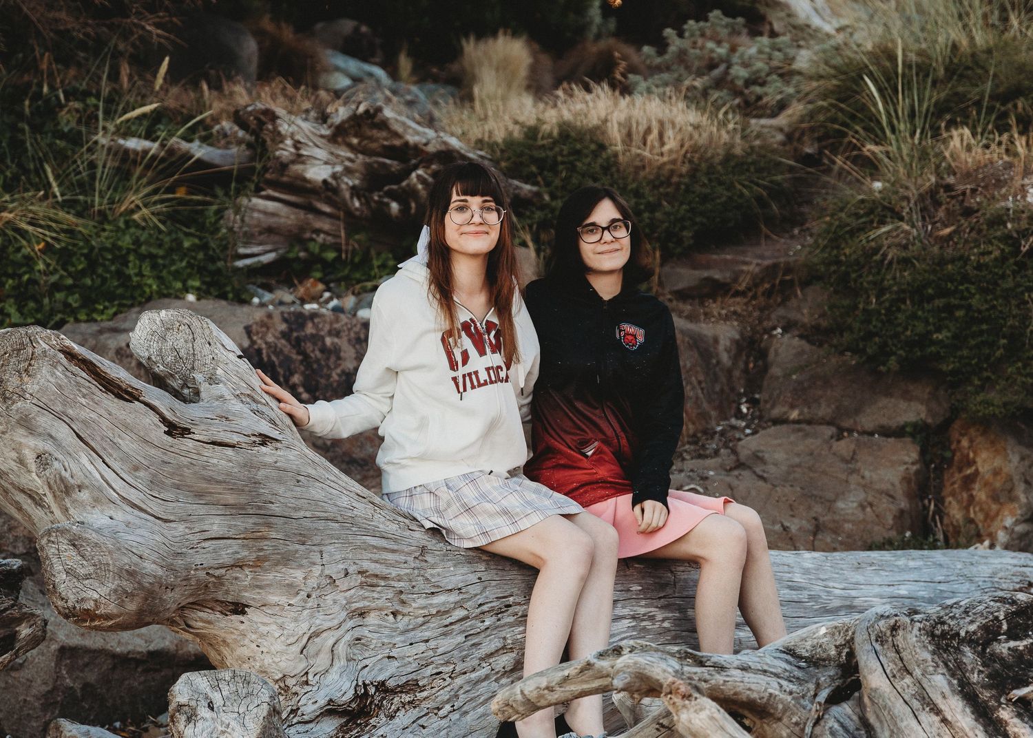 Two friends in contrasting black and white outfits sit together on driftwood at a beach during golden hour.