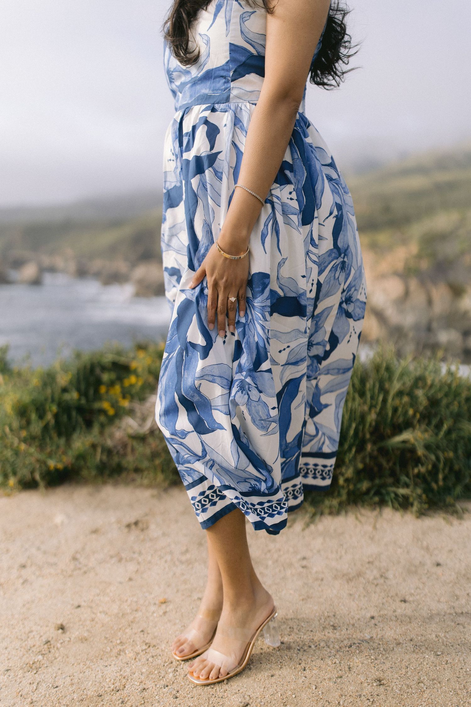 A person in a flowing blue and white floral dress stands barefoot on a coastal cliff overlooking the ocean.