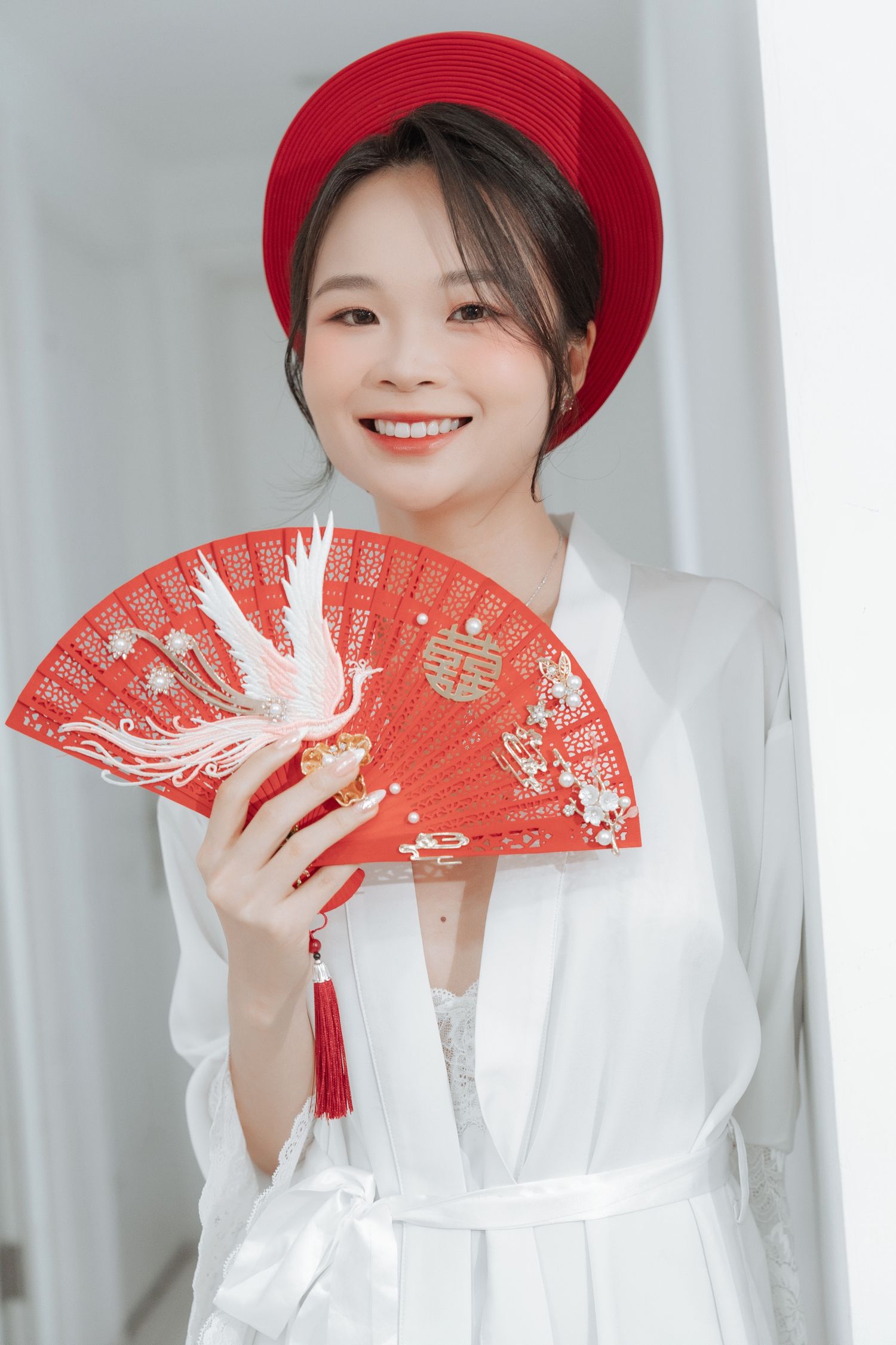 Bride in white robe and red hat displays ornate red fan with phoenix embroidery design.