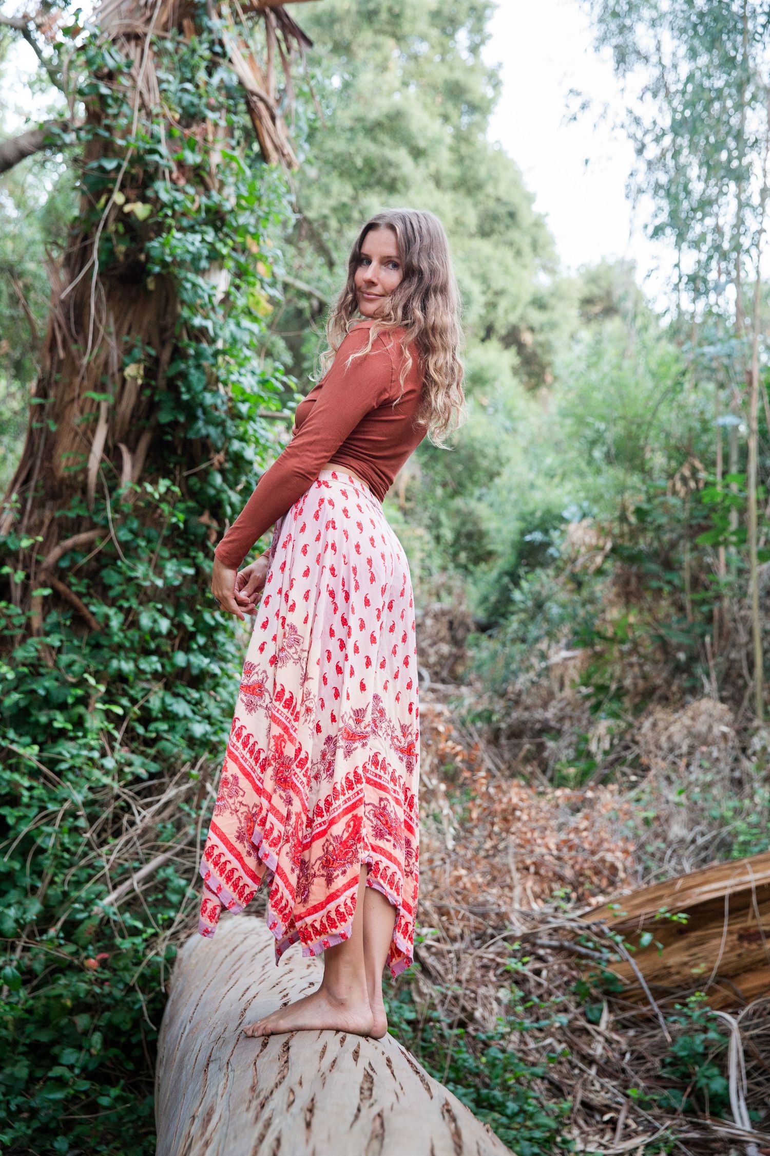 A woman in a red sweater and patterned skirt stands on a log in a lush forest setting.