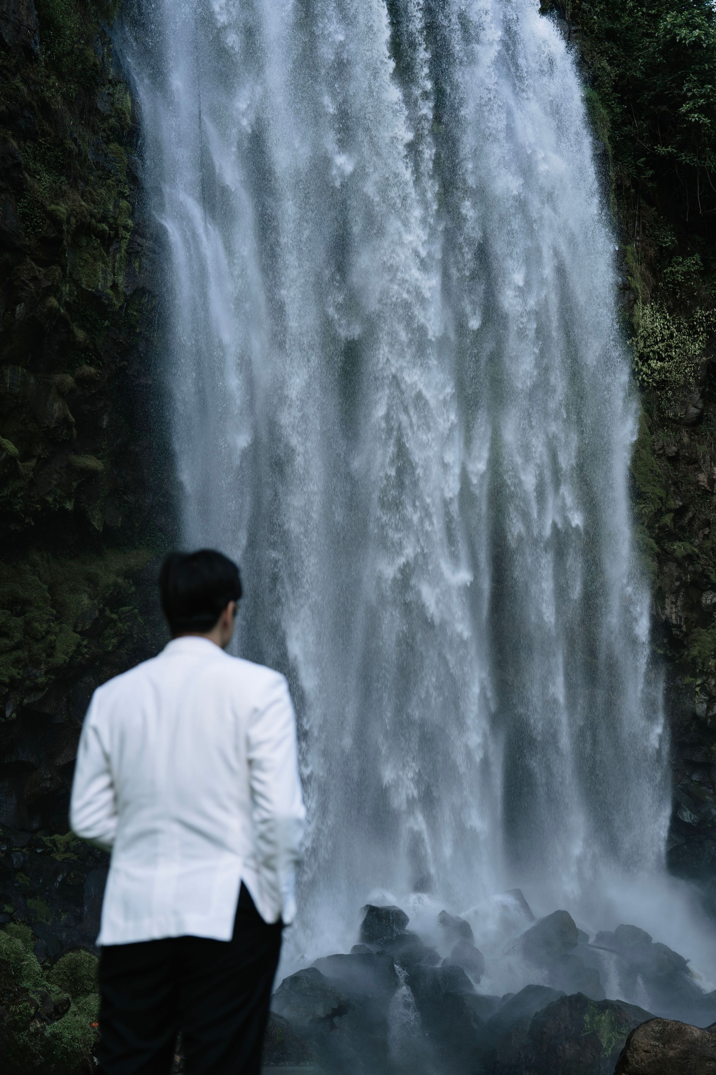 Groom in white tuxedo standing before a powerful cascading waterfall.