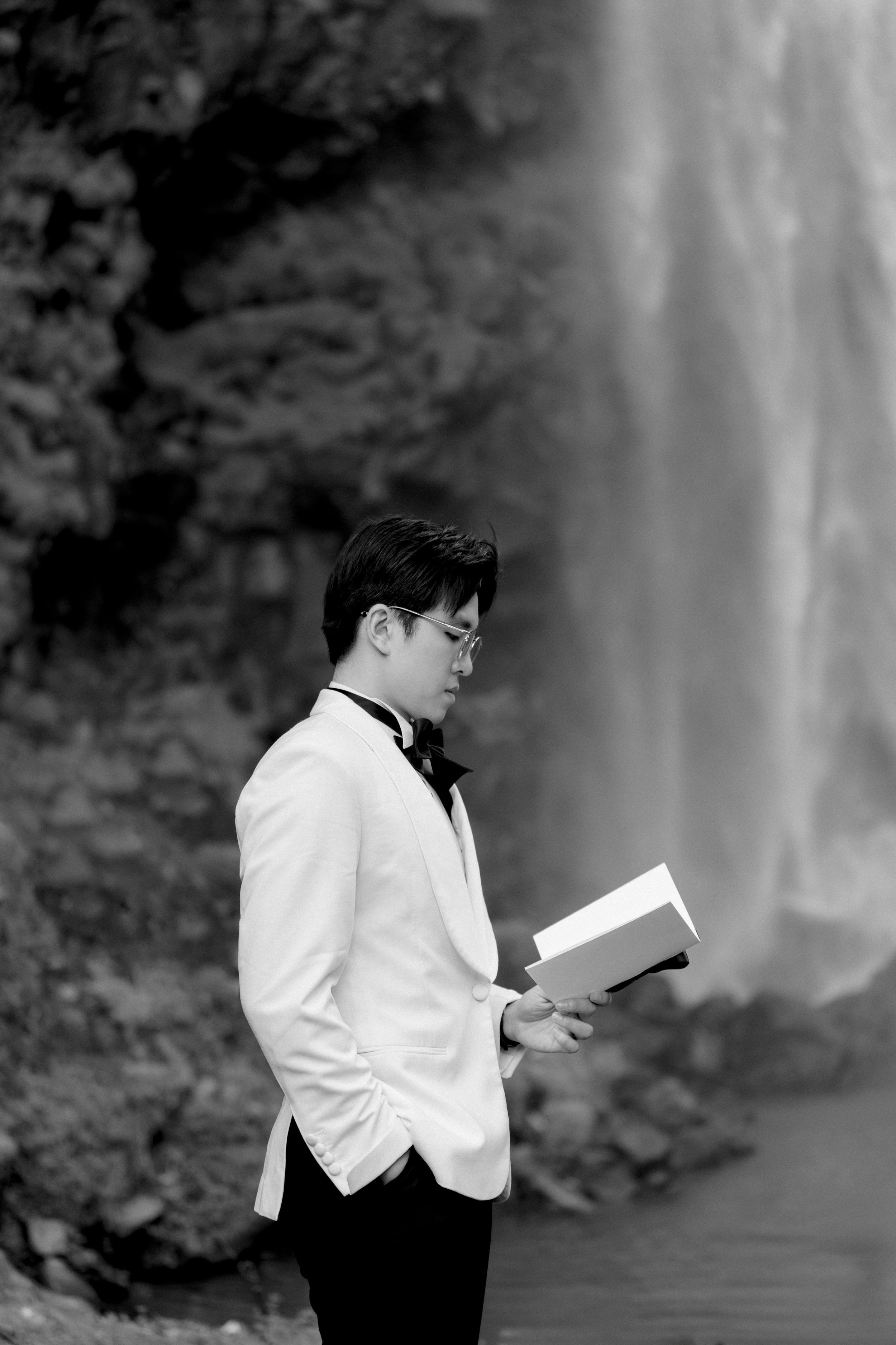 Black and white portrait of groom reading book with waterfall backdrop.