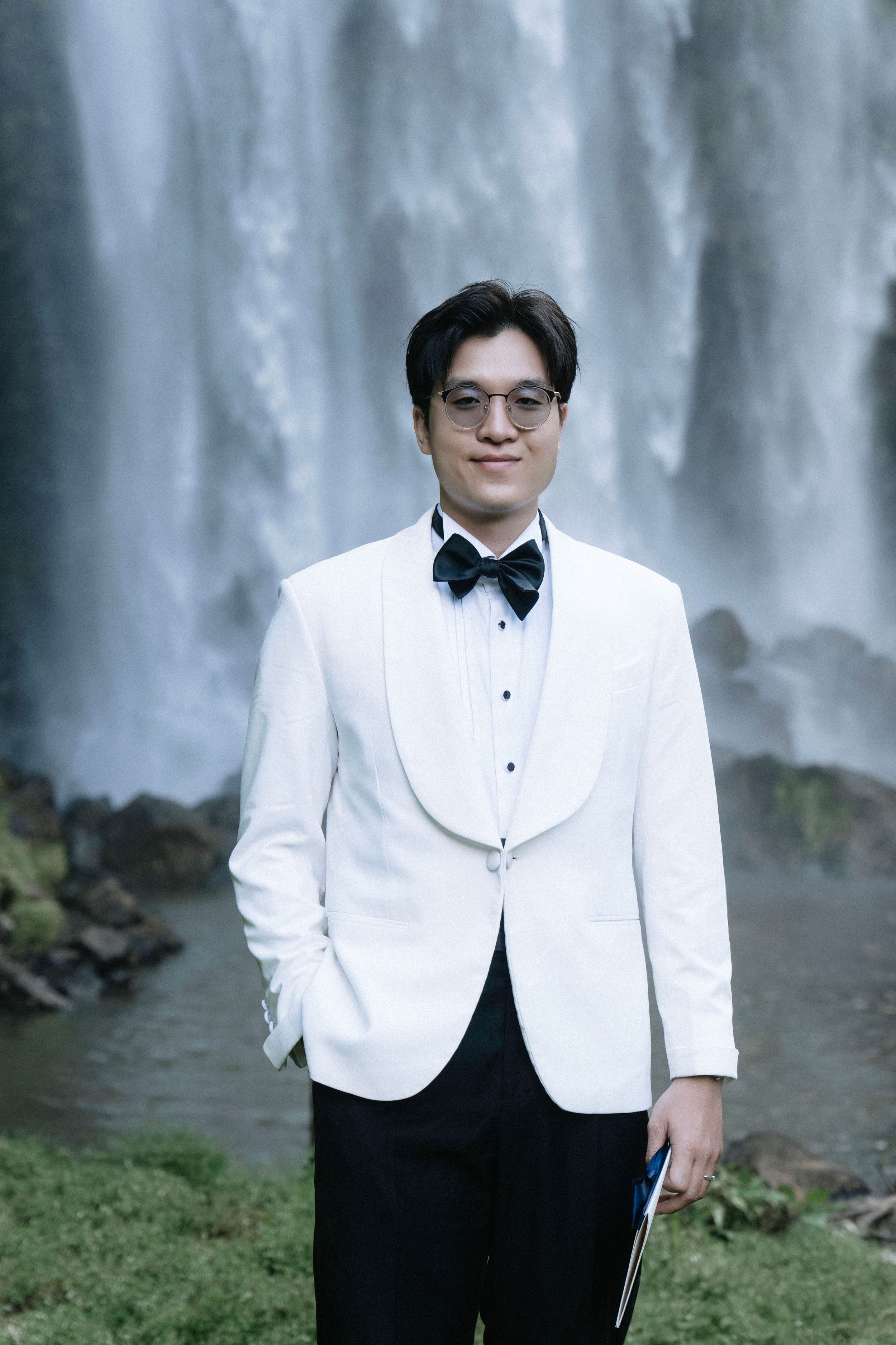 Man in white tuxedo jacket and black bow tie standing before a majestic waterfall.