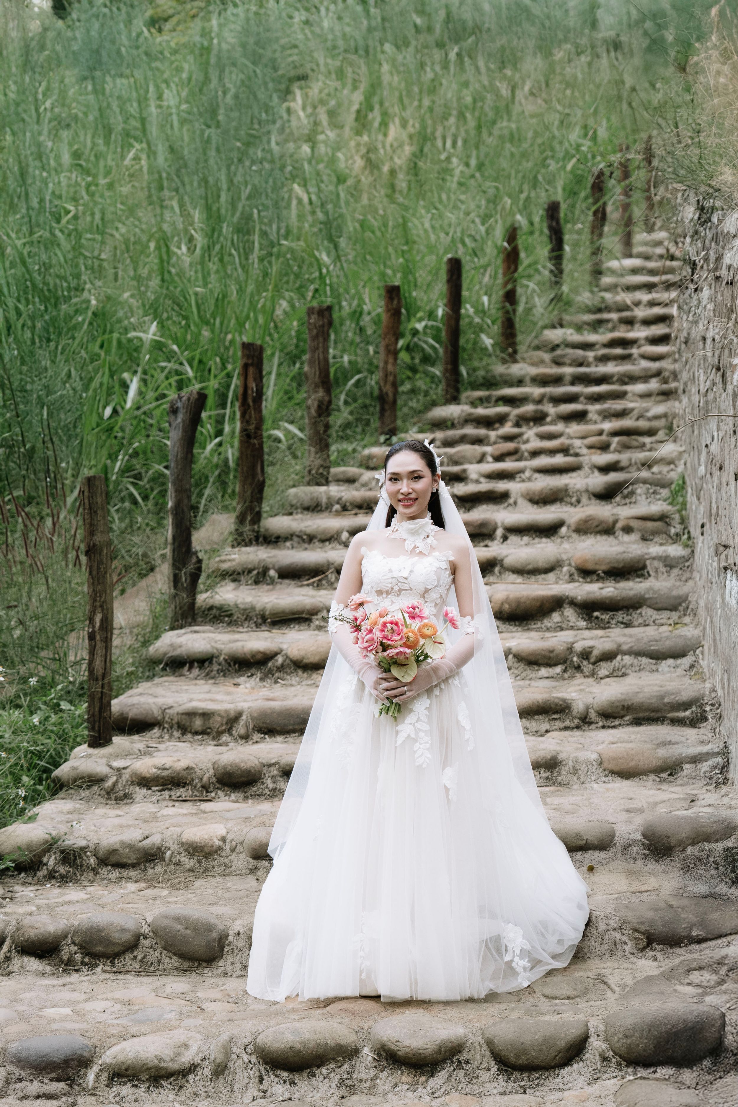Bride in white wedding gown holding pink bouquet on rustic stone staircase.