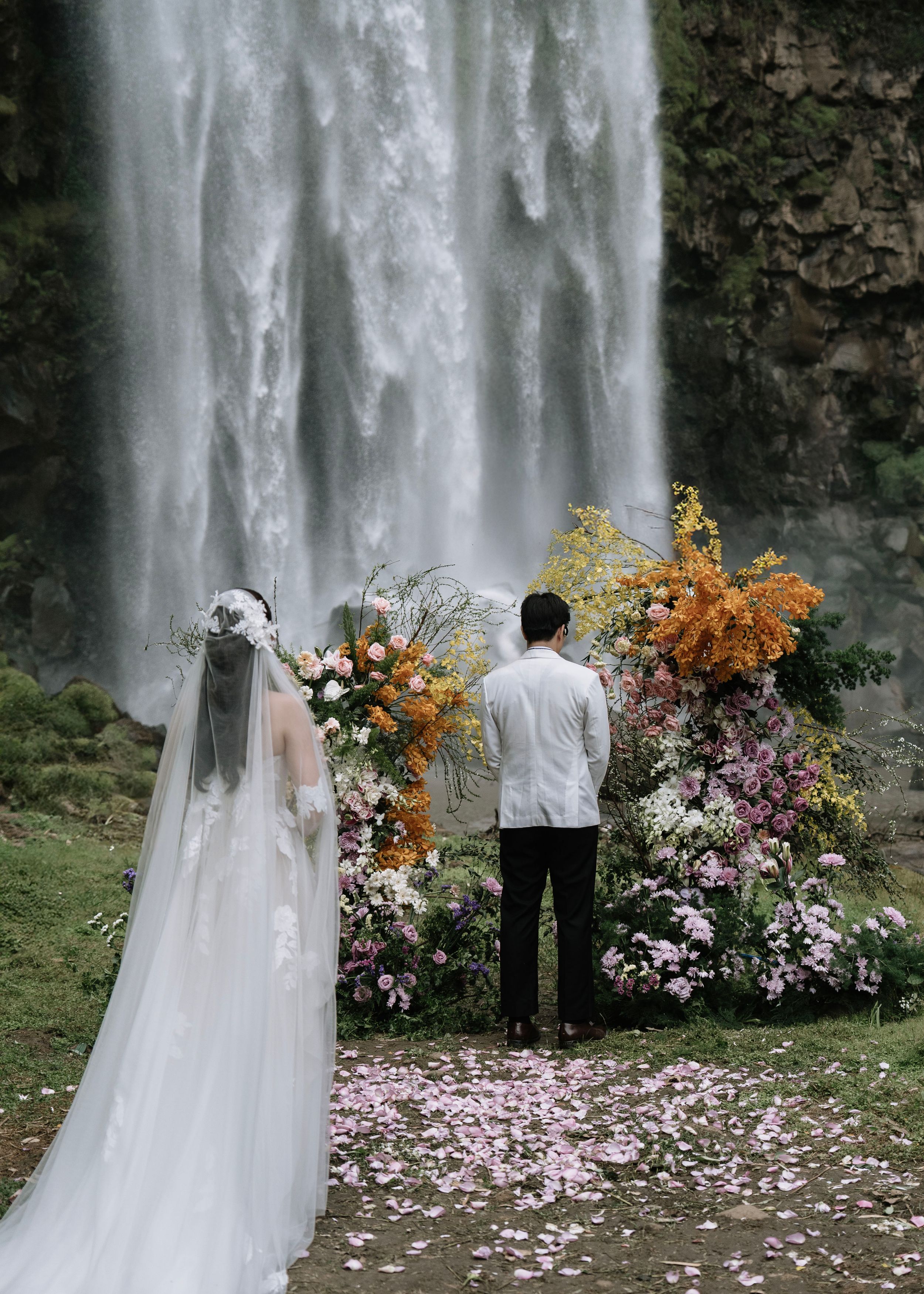 Couple exchanging vows at floral altar before a stunning waterfall backdrop.