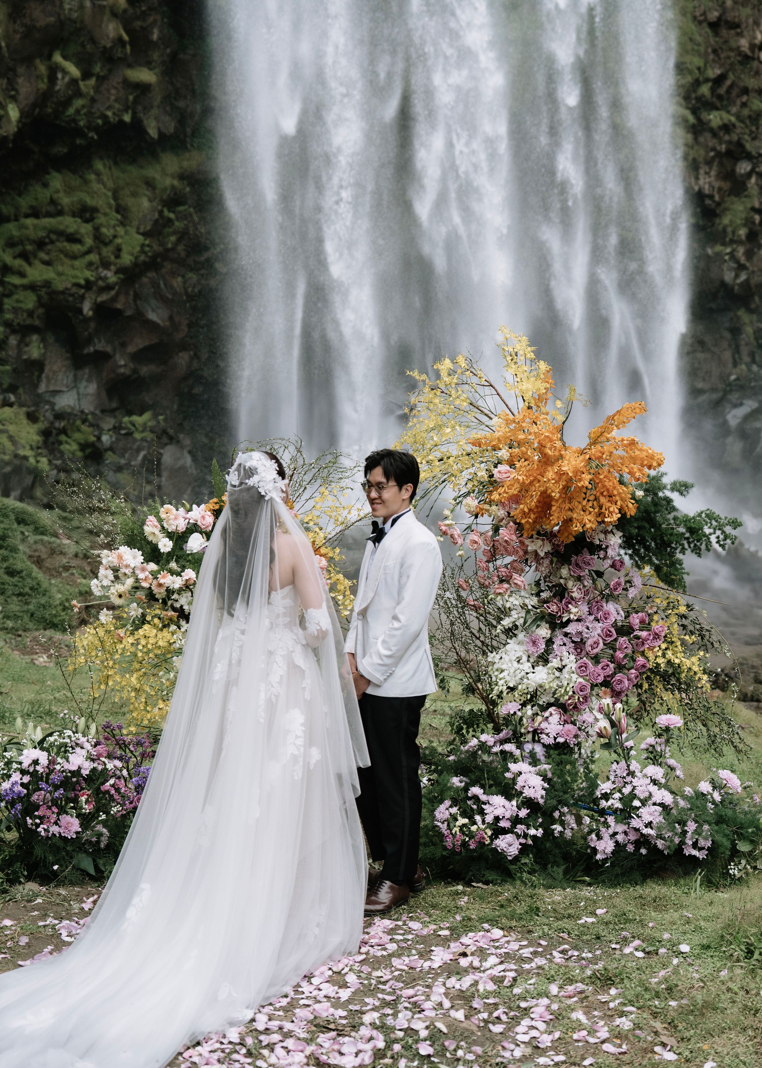 Bride and groom sharing intimate moment at flower-adorned waterfall ceremony.