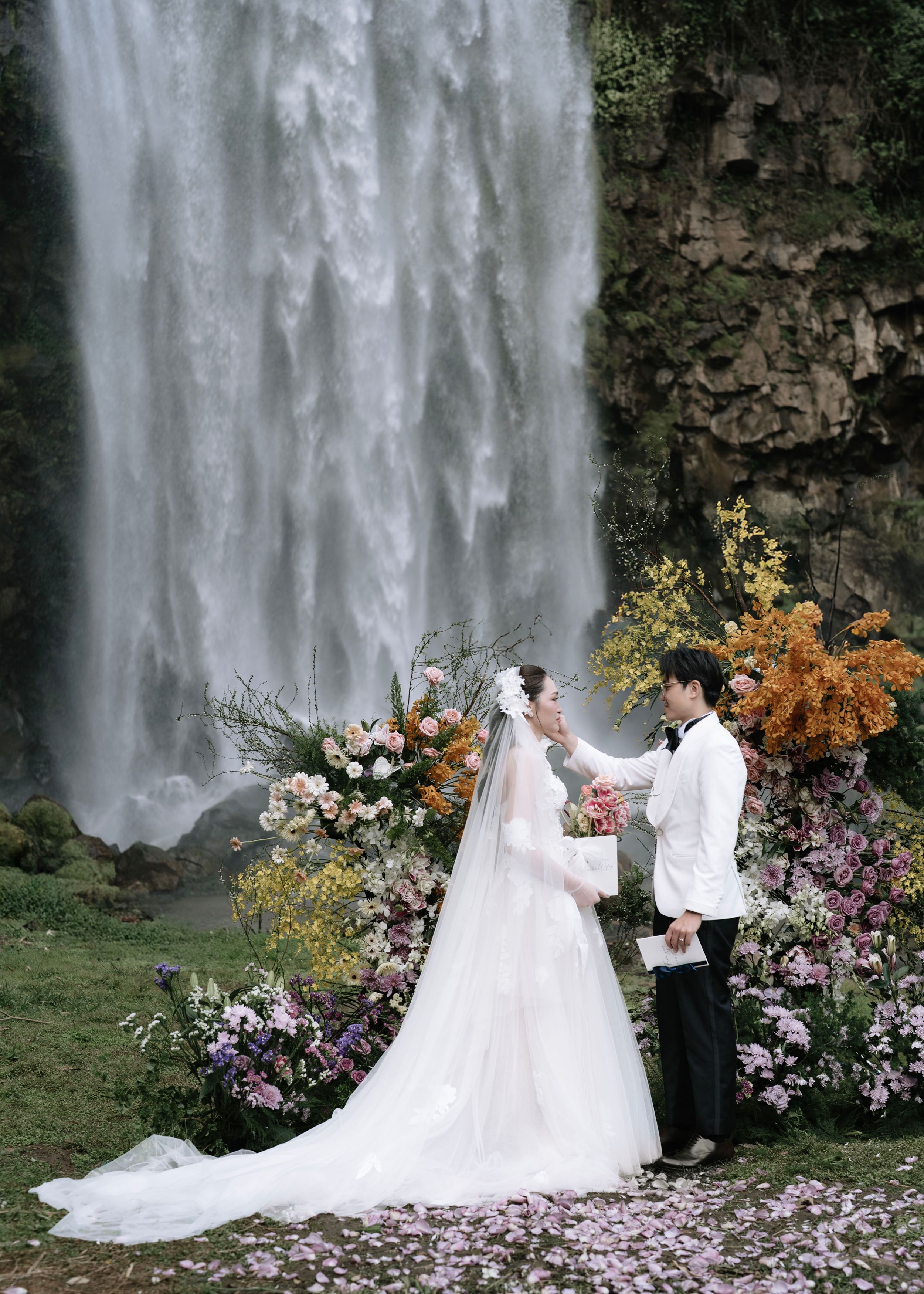 Bride and groom holding hands at colorful floral altar before a waterfall.