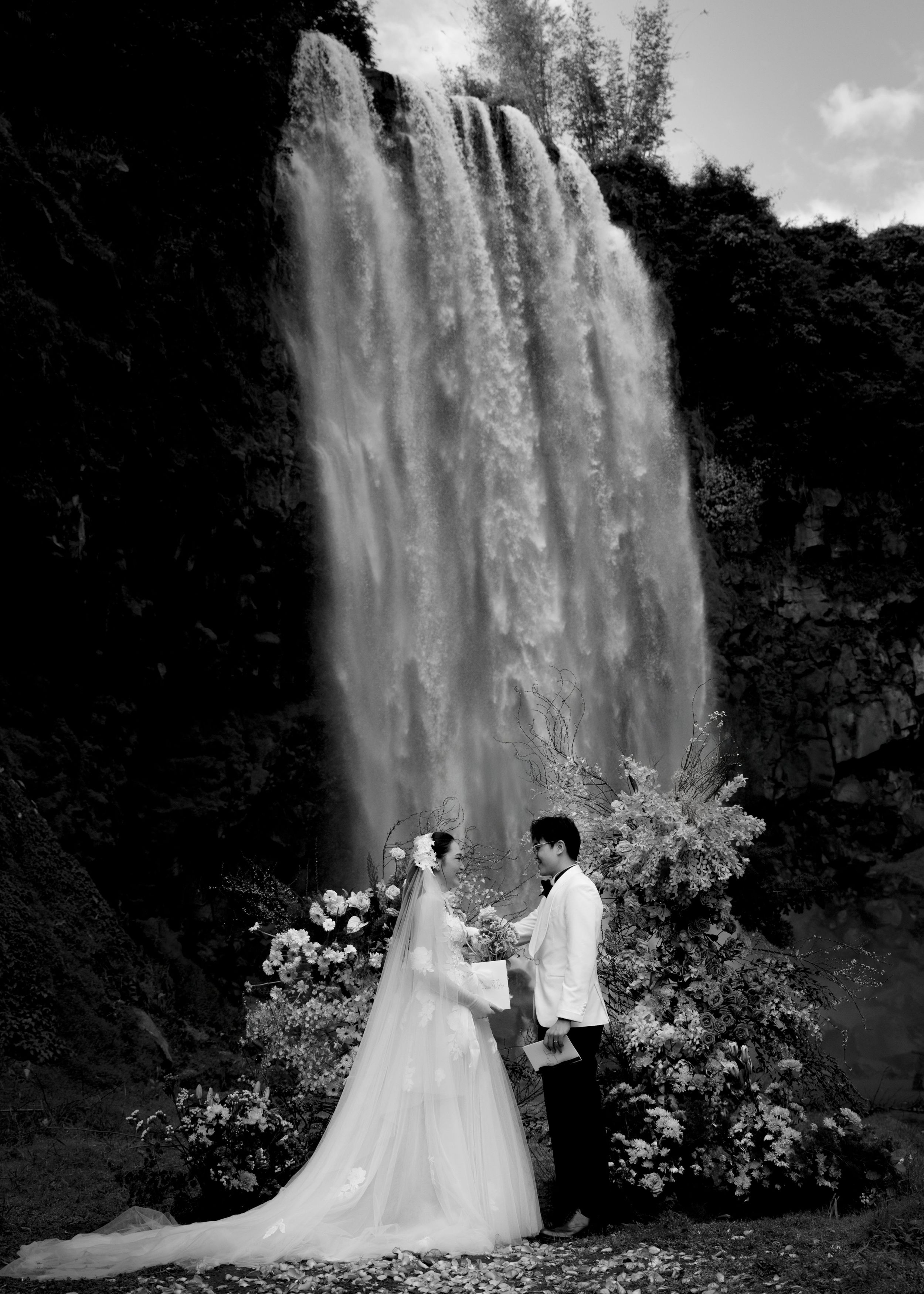 Black and white photo of couple exchanging vows at floral waterfall ceremony.