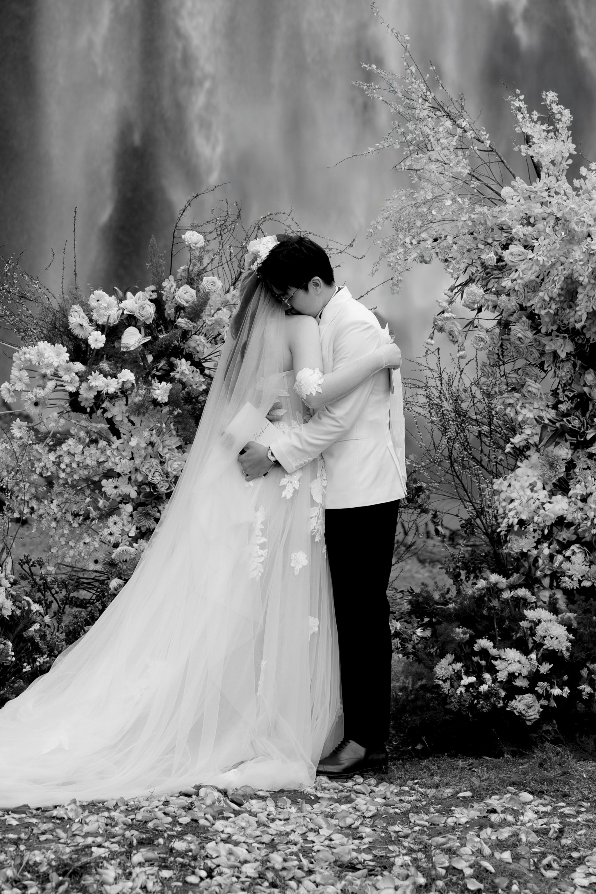 Black and white photo of bride and groom embracing at floral waterfall altar.