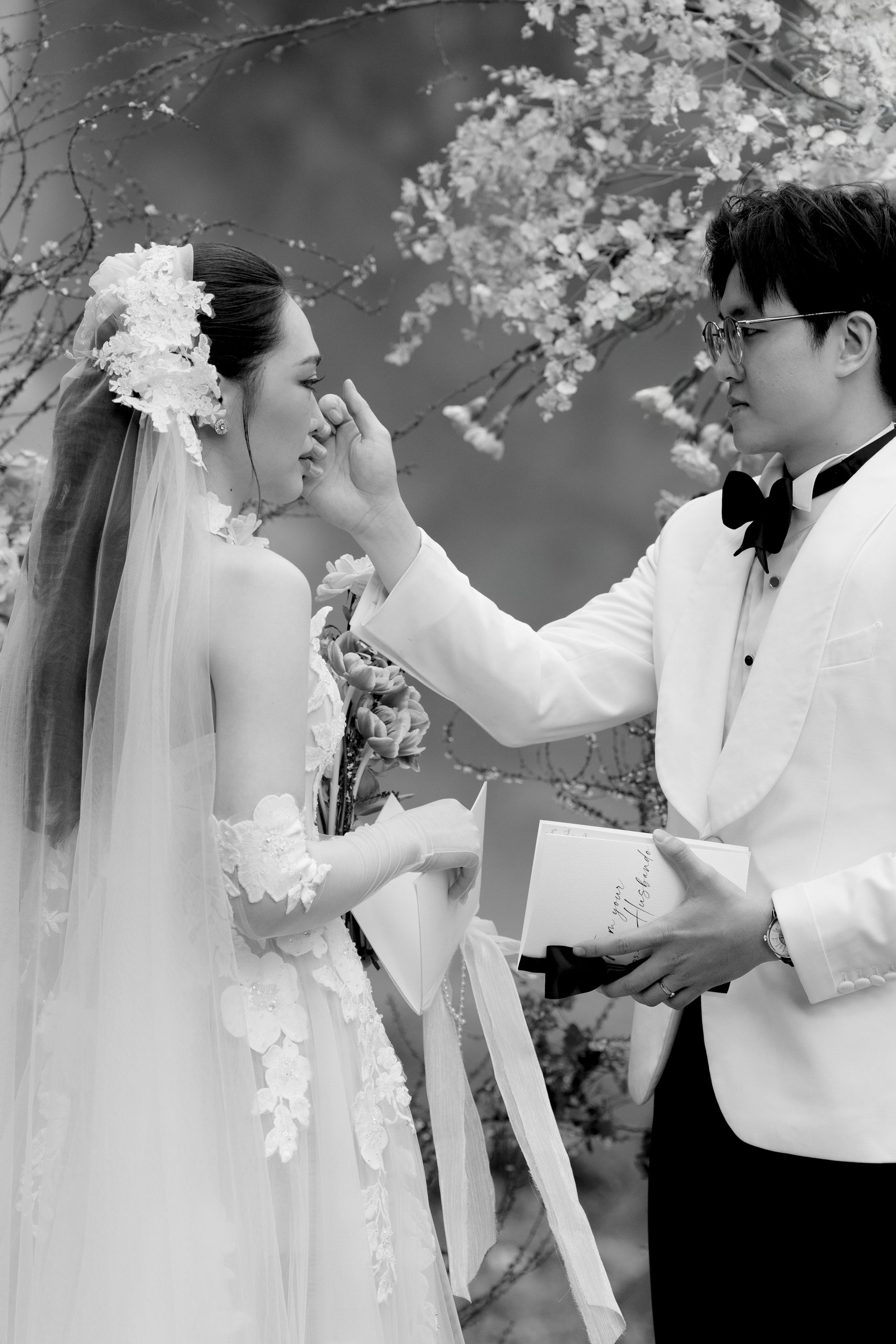 Groom tenderly wiping bride's tears during emotional outdoor wedding ceremony with cherry blossoms.