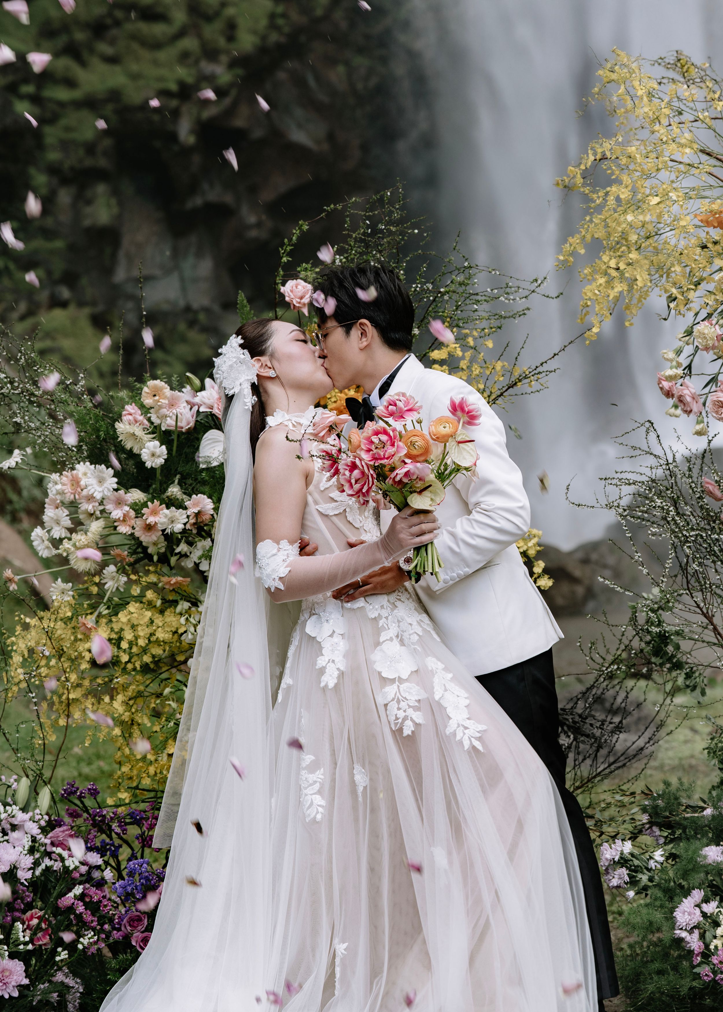 Couple sharing romantic kiss surrounded by colorful flowers with waterfall backdrop on wedding day.