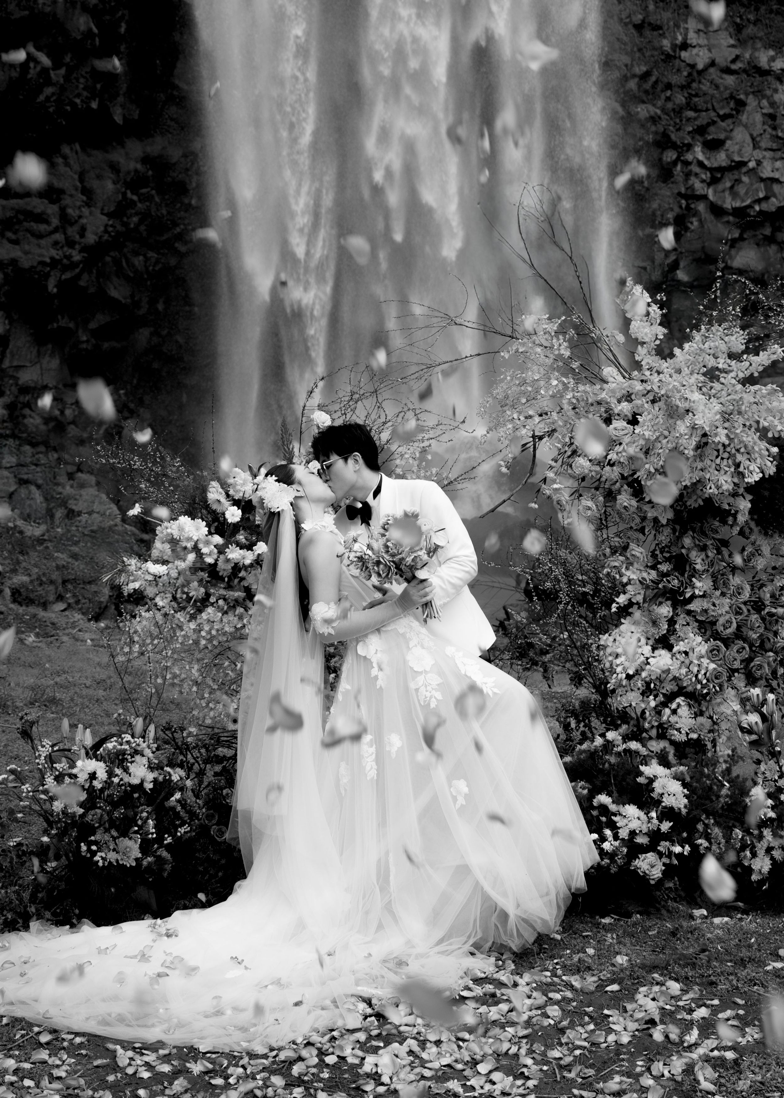Black and white photo of bride and groom kissing near waterfall with floral arrangements.