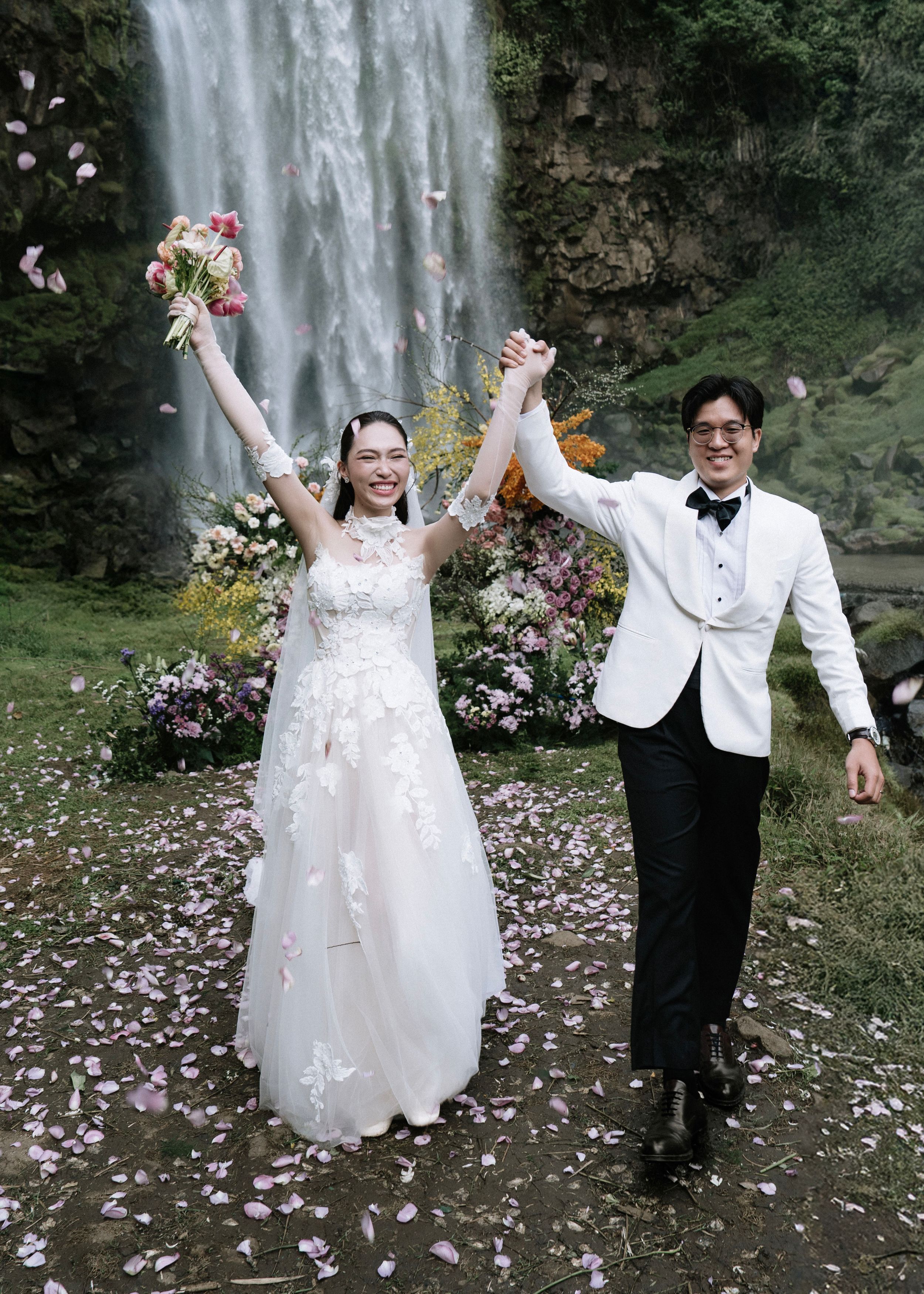 Joyful newlyweds celebrating with raised bouquet and held hands before a stunning waterfall backdrop.